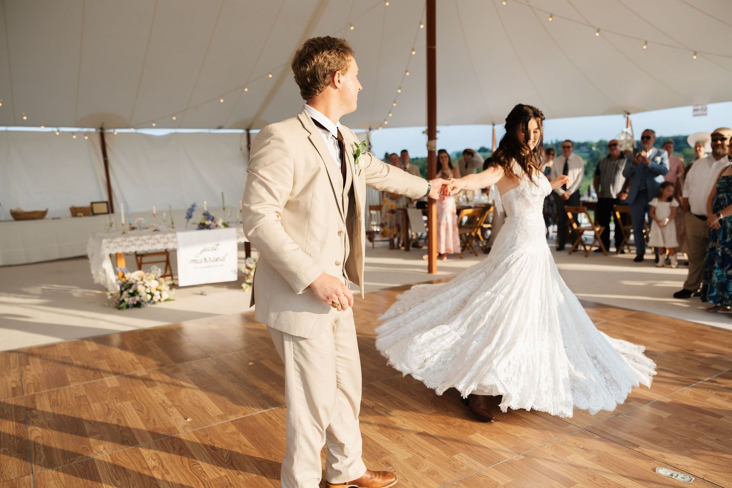 Bride and groom share first dance under white tent at outdoor wedding reception on wooden dance floor.