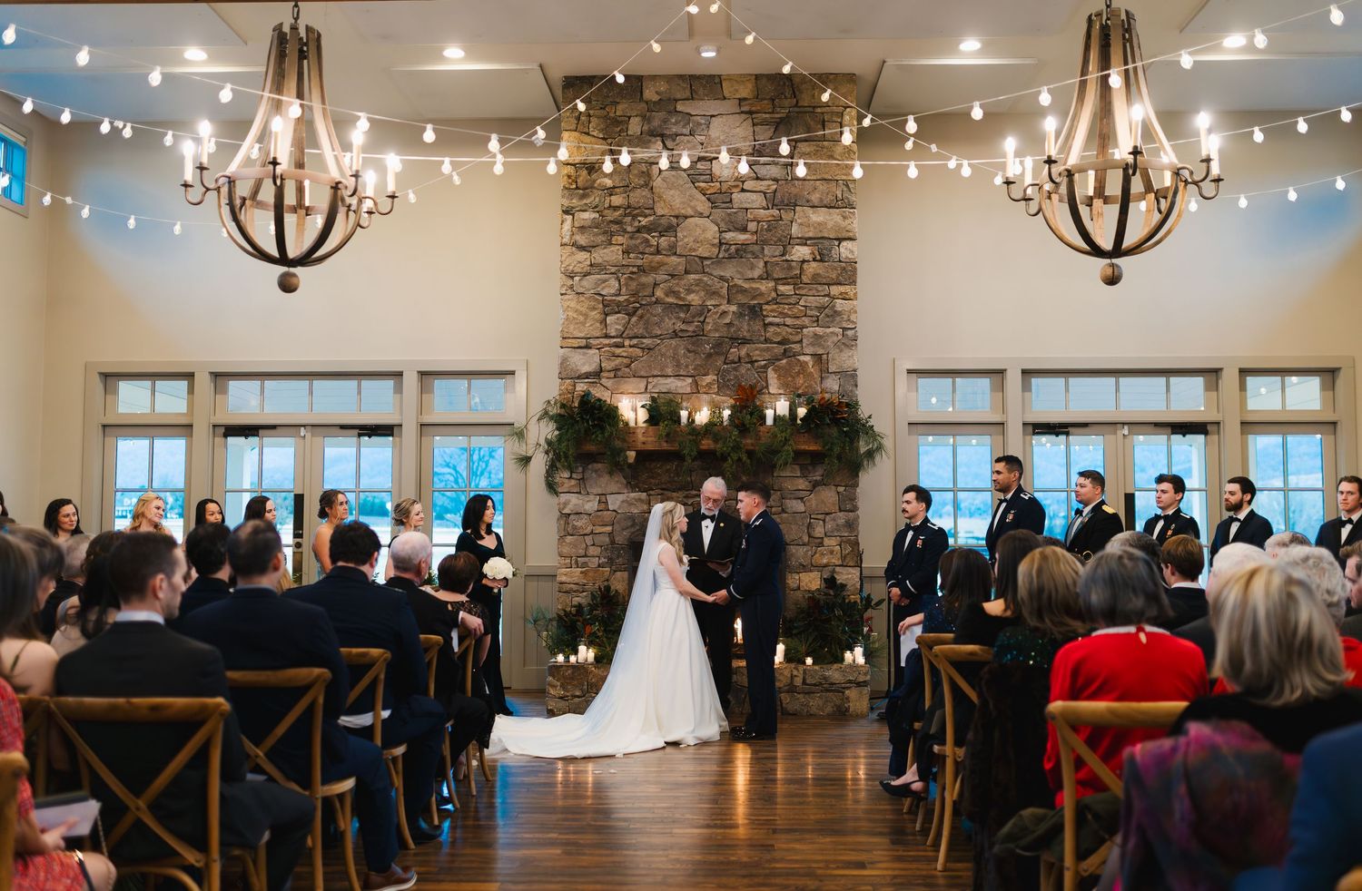 Elegant indoor wedding ceremony with stone fireplace, chandeliers, and guests seated in wooden chairs.