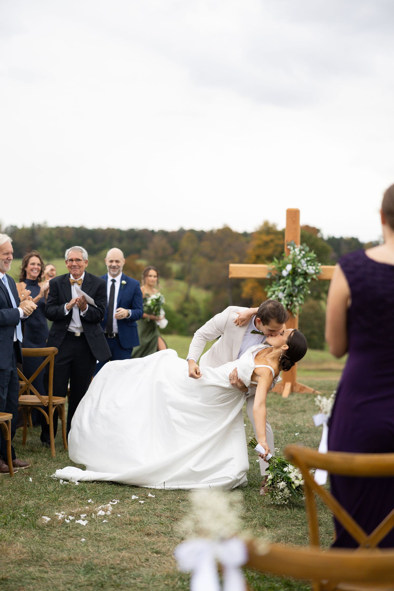 Groom dips bride for romantic kiss at outdoor ceremony while guests cheer and applaud.