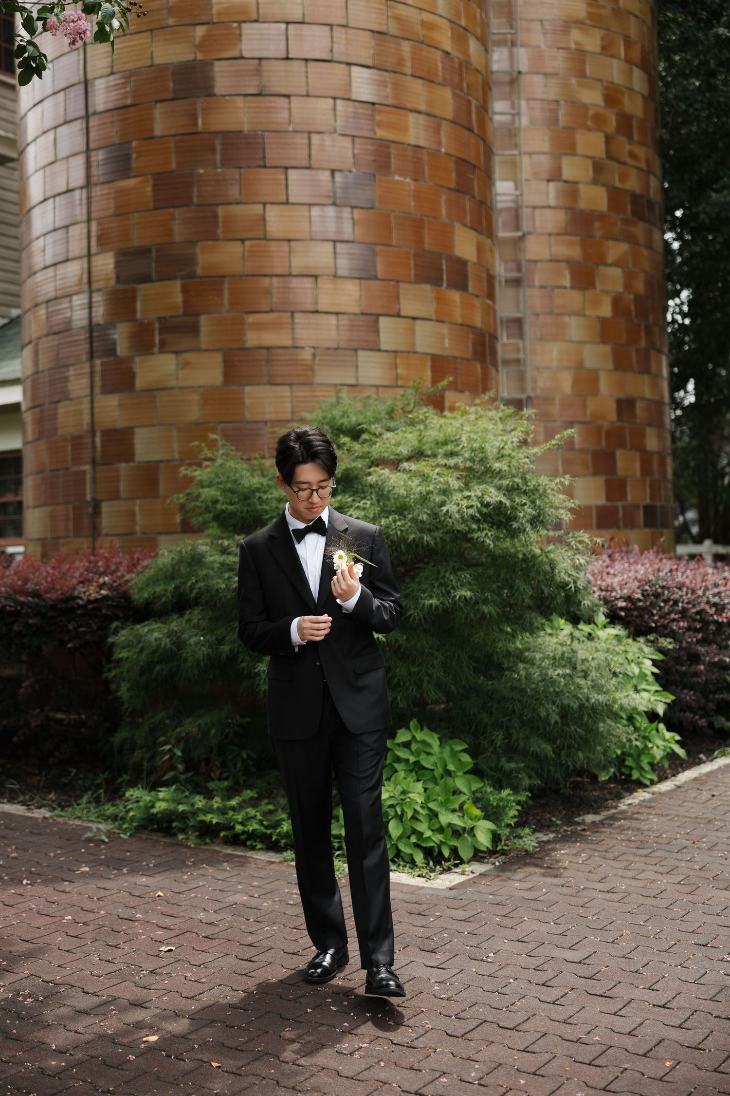 A groom in a black tuxedo adjusts his boutonniere while standing on a brick path near lush greenery.