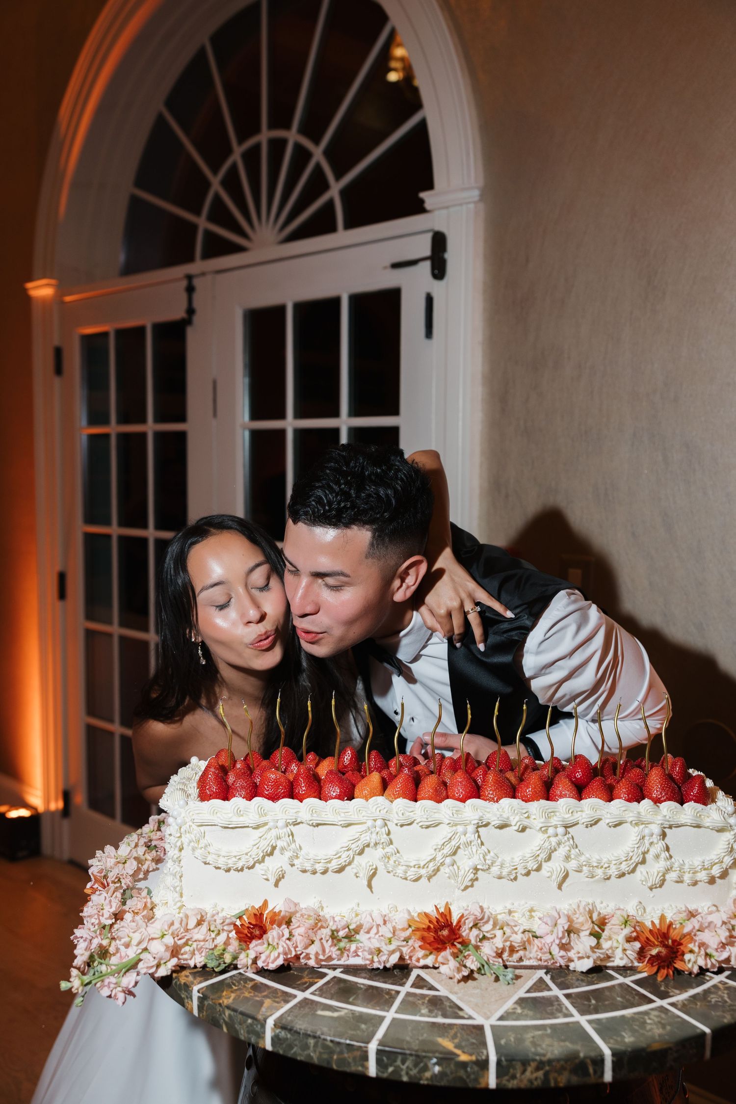 Bride and groom share a sweet moment leaning in to kiss over white wedding cake topped with fresh strawberries.