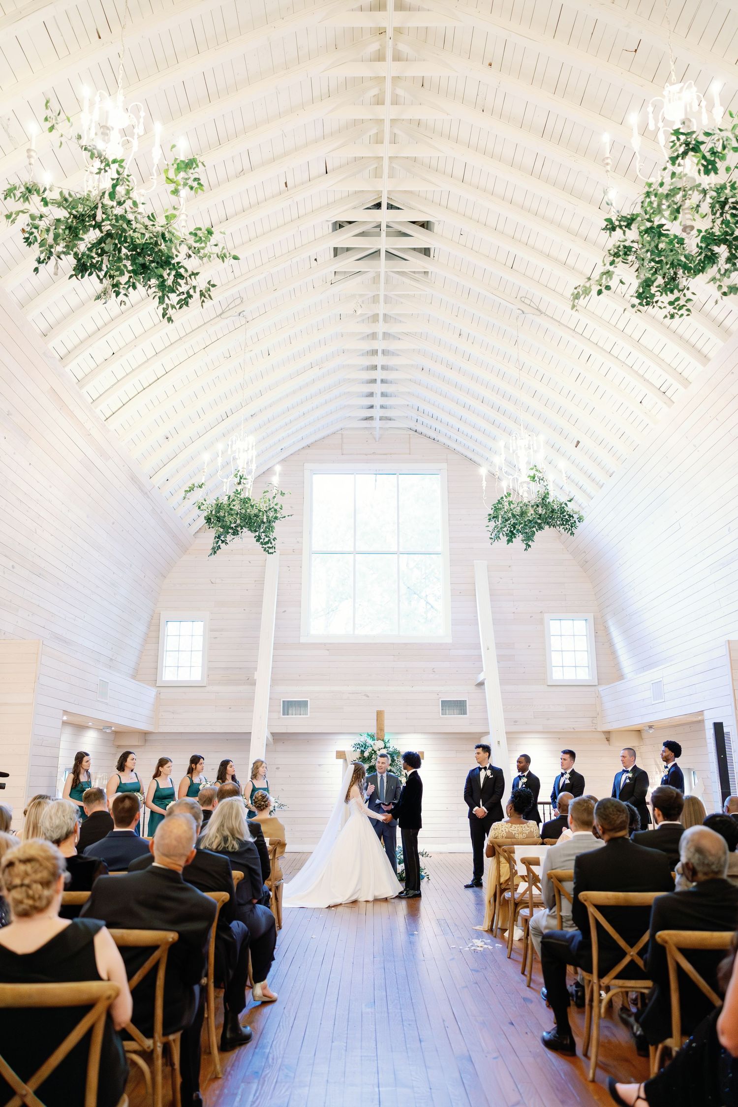 Wide-angle view of elegant white barn wedding ceremony with greenery chandeliers and seated guests.