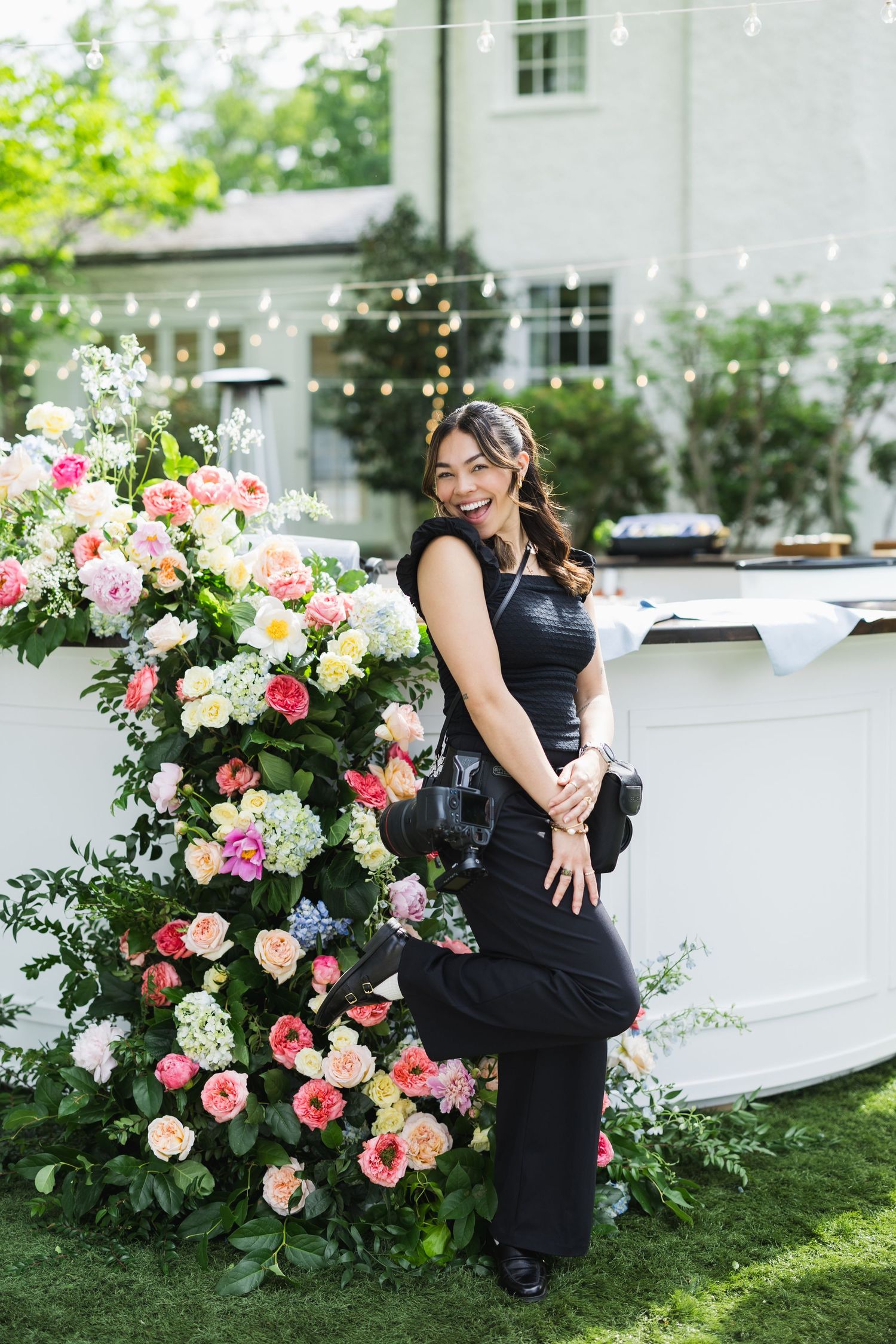Smiling photographer in black outfit posing beside colorful floral arrangement at outdoor event venue.
