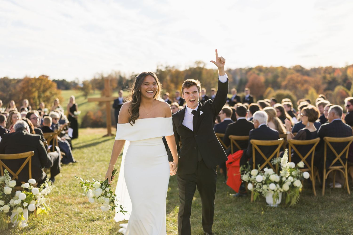Smiling newlyweds celebrating recessional at outdoor autumn wedding ceremony with guests seated on hillside.