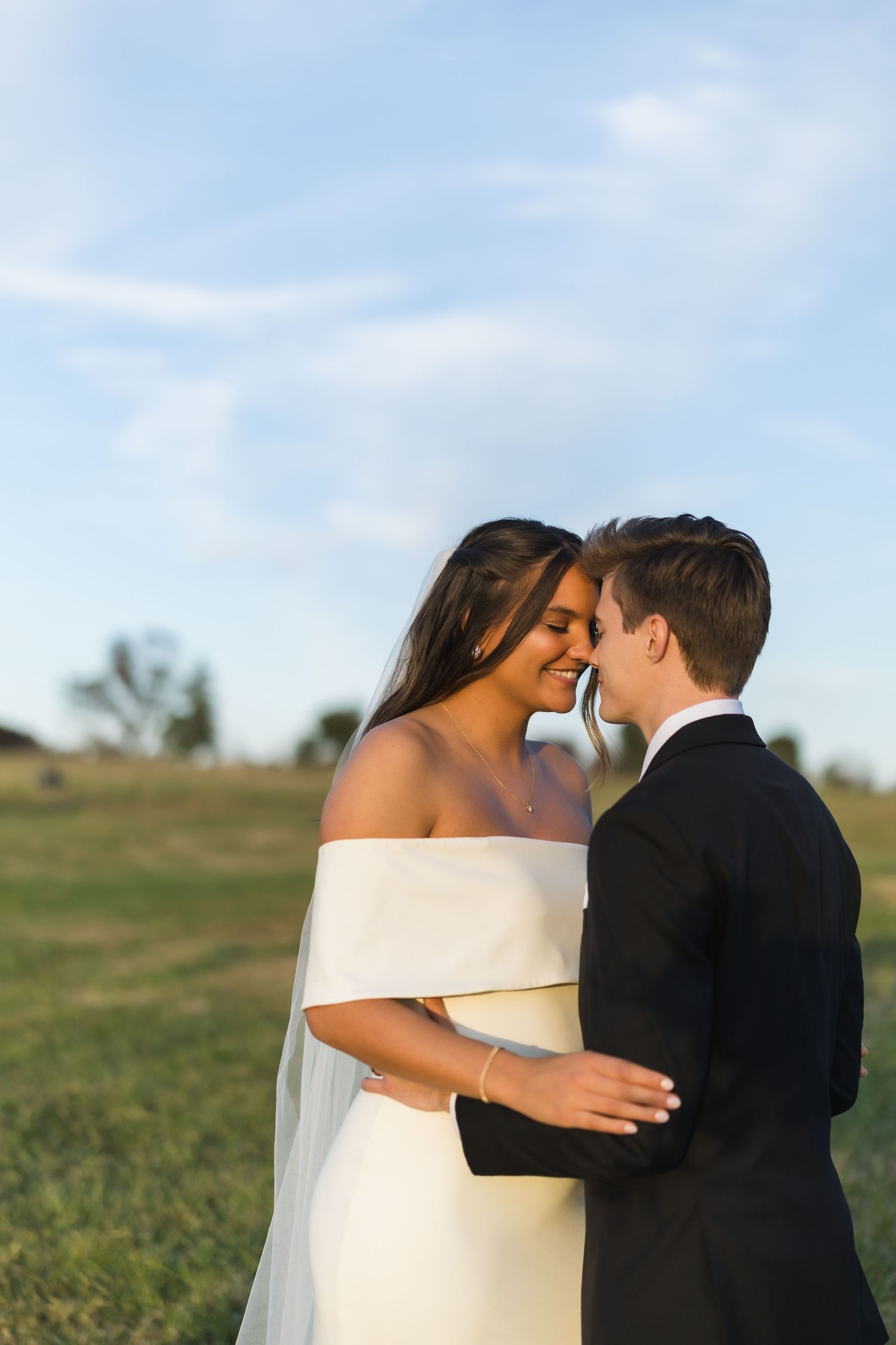 Bride and groom share an intimate moment, nearly kissing in a sunlit green field on their wedding day.