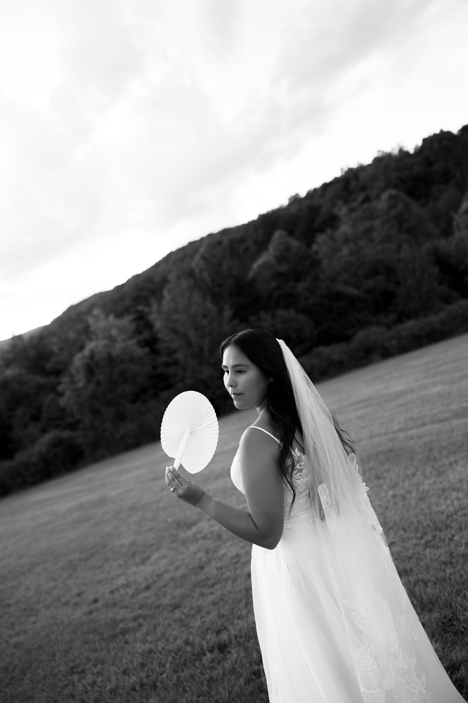 Black and white photo of a bride in a white gown and veil holding a fan in an open field with trees.
