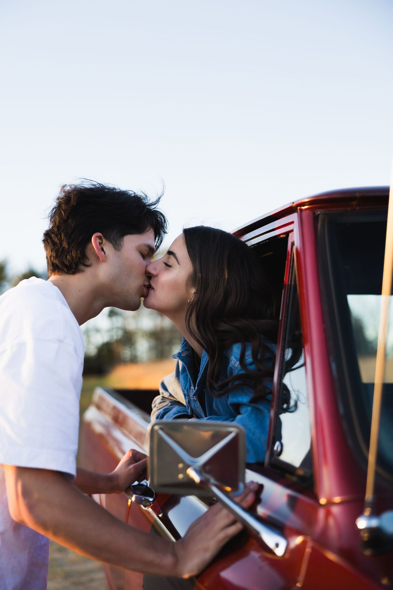 Romantic couple sharing a kiss through the window of a classic red pickup truck at dusk in the countryside.