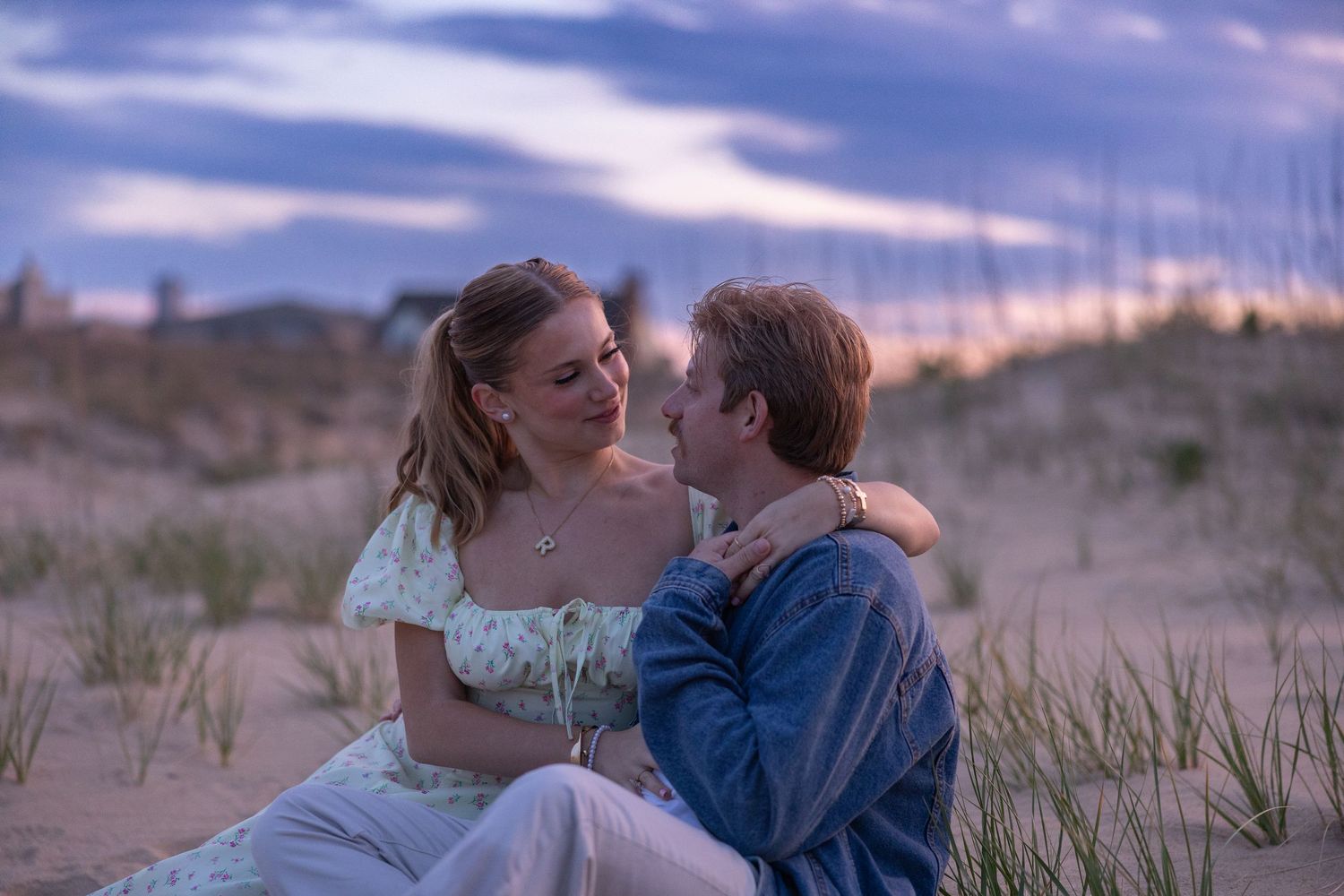 Couple gazes lovingly at each other while sitting on sand dunes at twilight sunset.