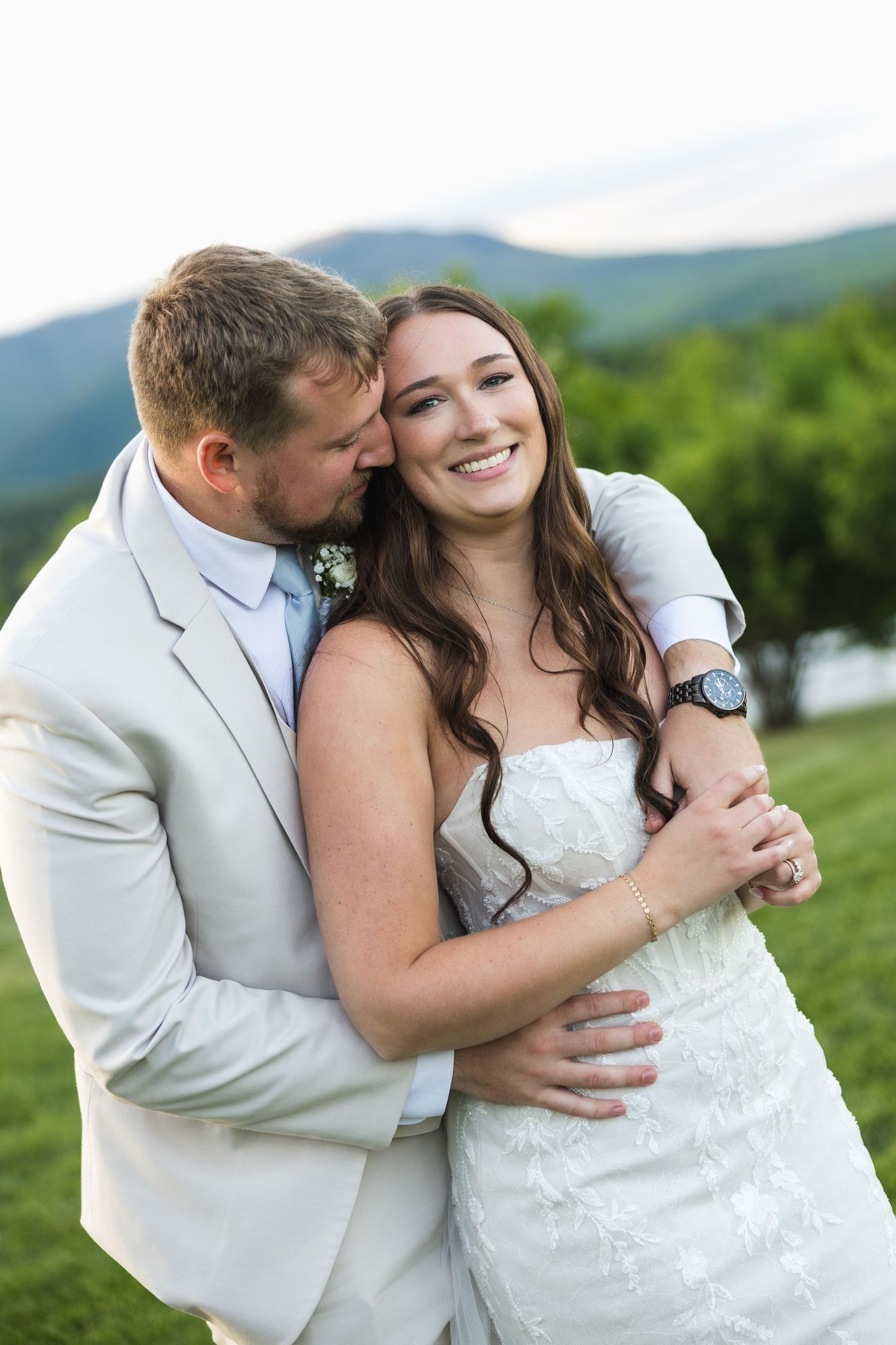 Bride and groom embracing outdoors with mountain views, groom in light suit kissing bride's cheek as she smiles.
