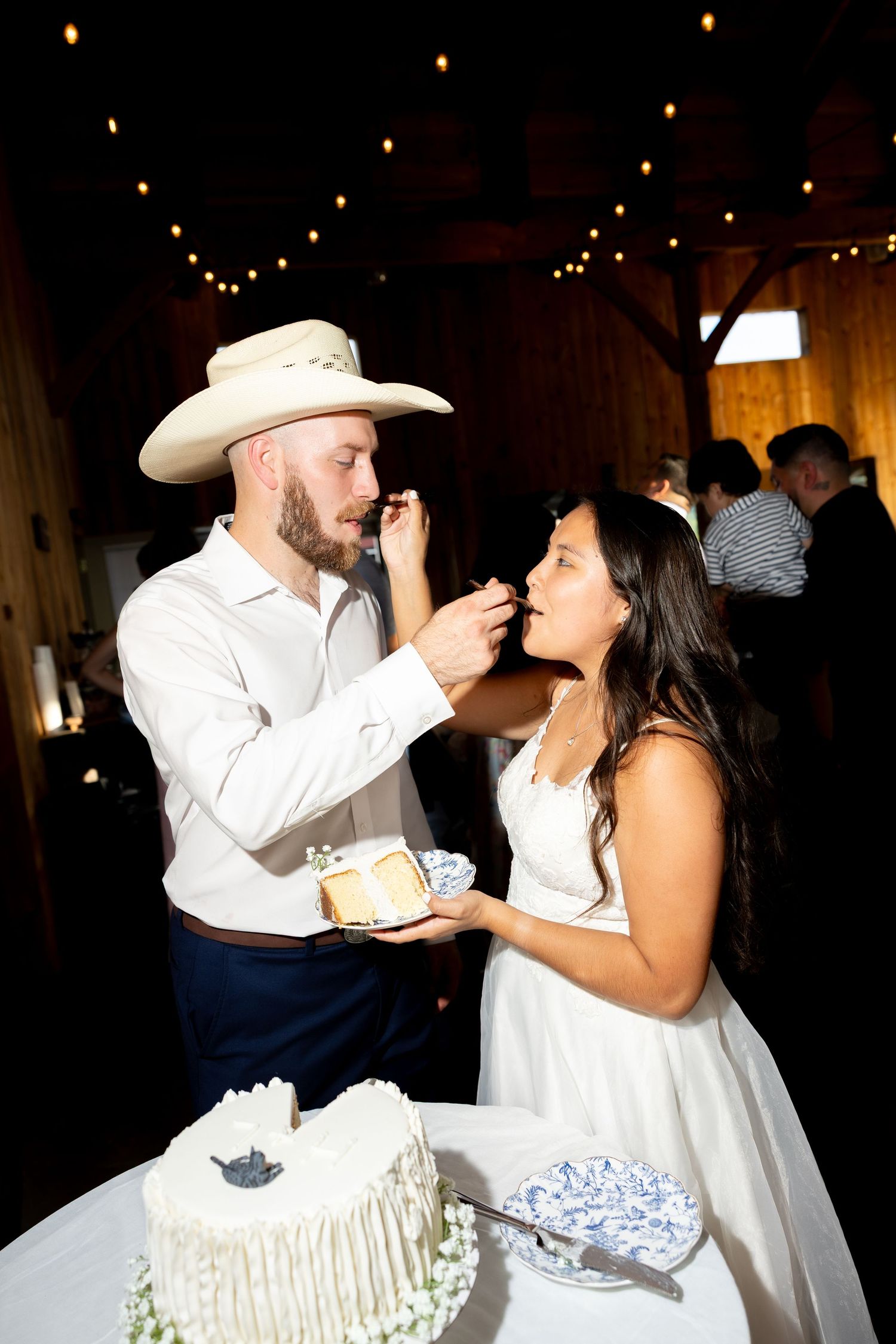 Bride and groom share wedding cake at rustic barn reception, groom wearing cowboy hat feeds bride a bite.