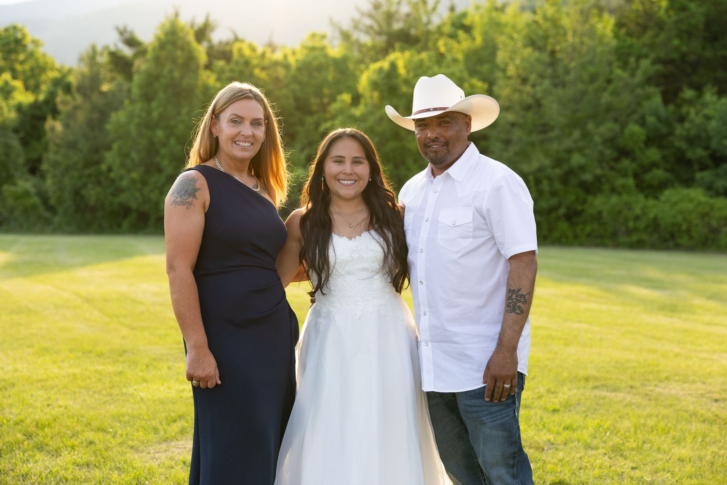 Family portrait with woman in navy dress, girl in white gown, and man in cowboy hat outdoors.