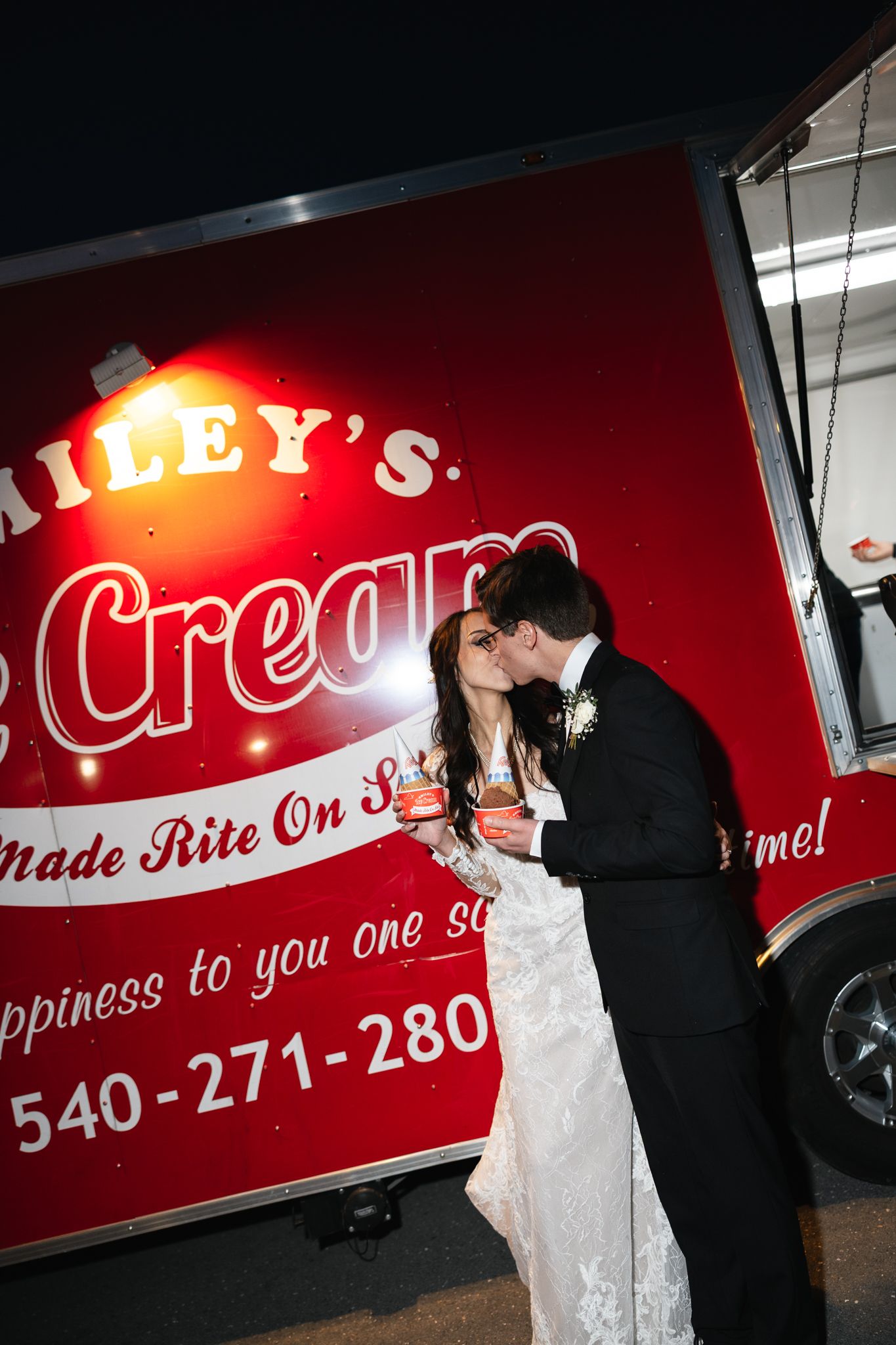 Newlyweds kissing while holding ice cream cups in front of a red Hailey's Ice Cream truck at night.