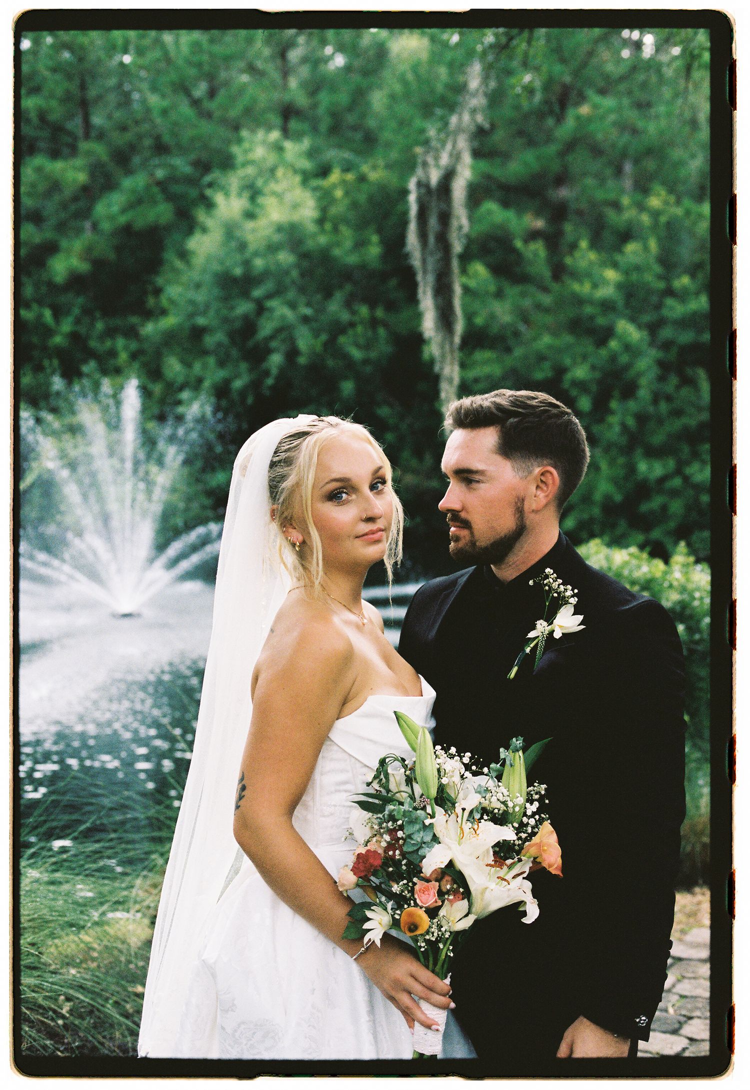 Bride in white strapless gown with veil holds bouquet beside groom in black suit near fountain.