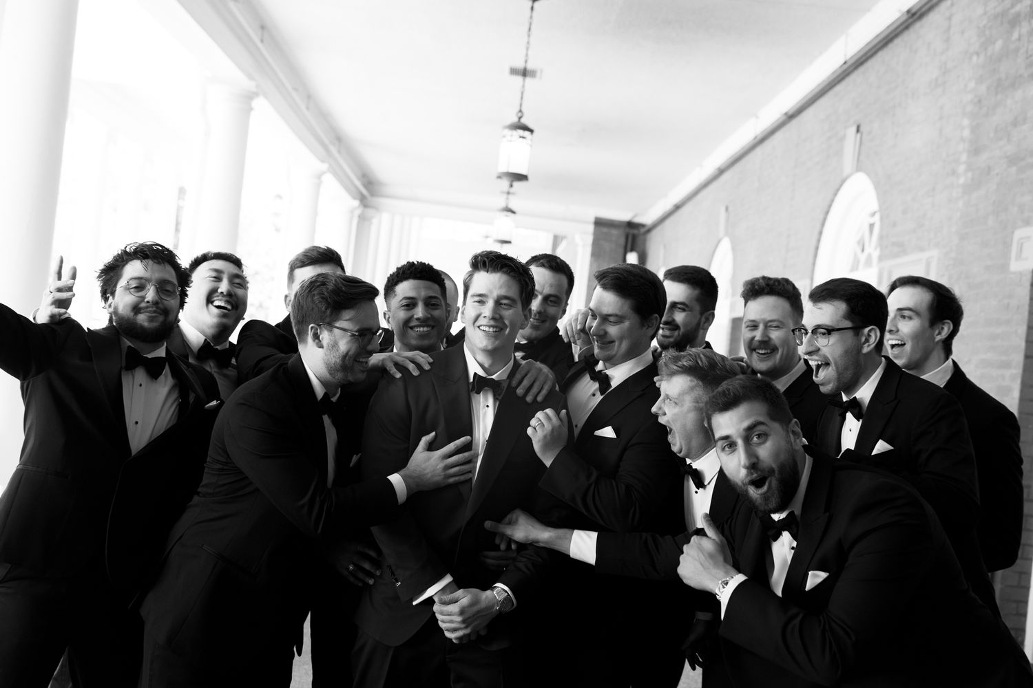 Black and white photo of a large groomsmen group in tuxedos laughing and posing together in an elegant hallway.
