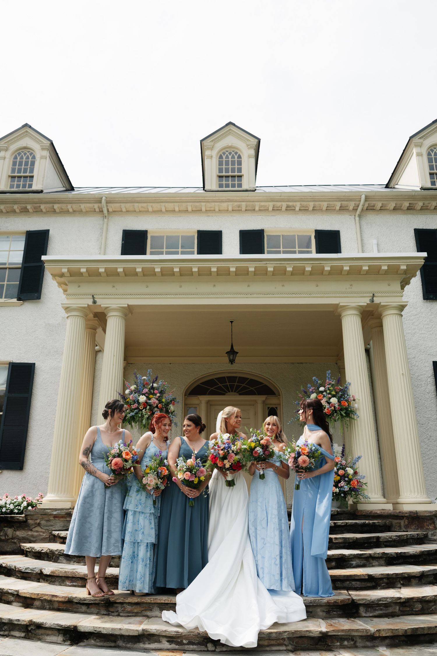 Bride in white gown with bridesmaids in blue dresses holding colorful bouquets on steps of elegant white mansion.