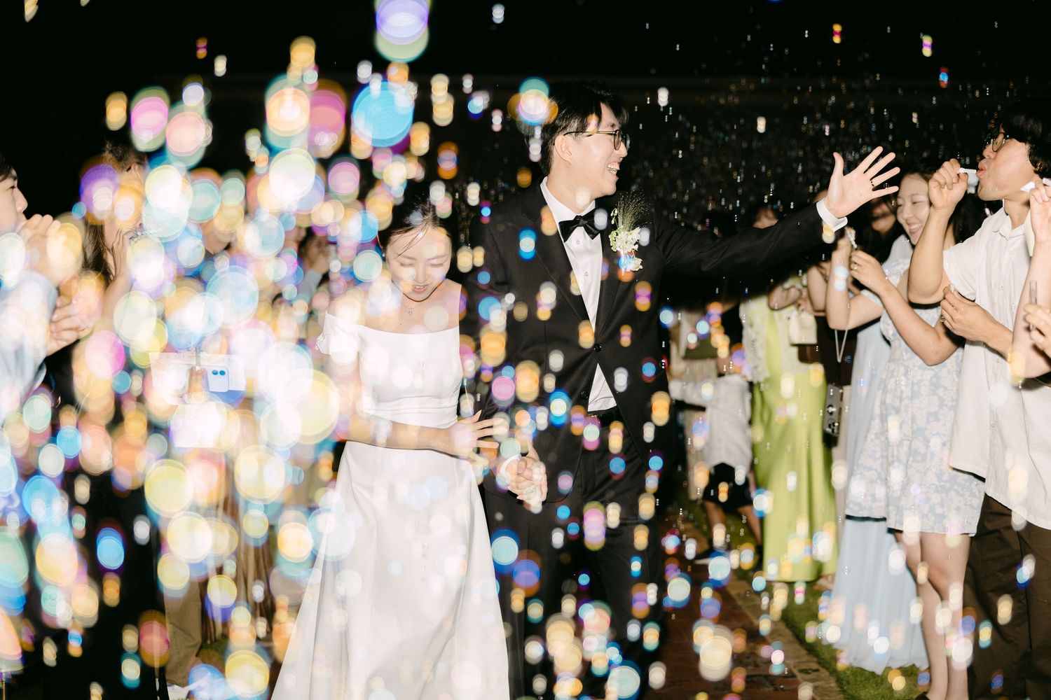Couple in wedding attire walks through colorful bokeh lights while guests celebrate at an elegant nighttime reception.