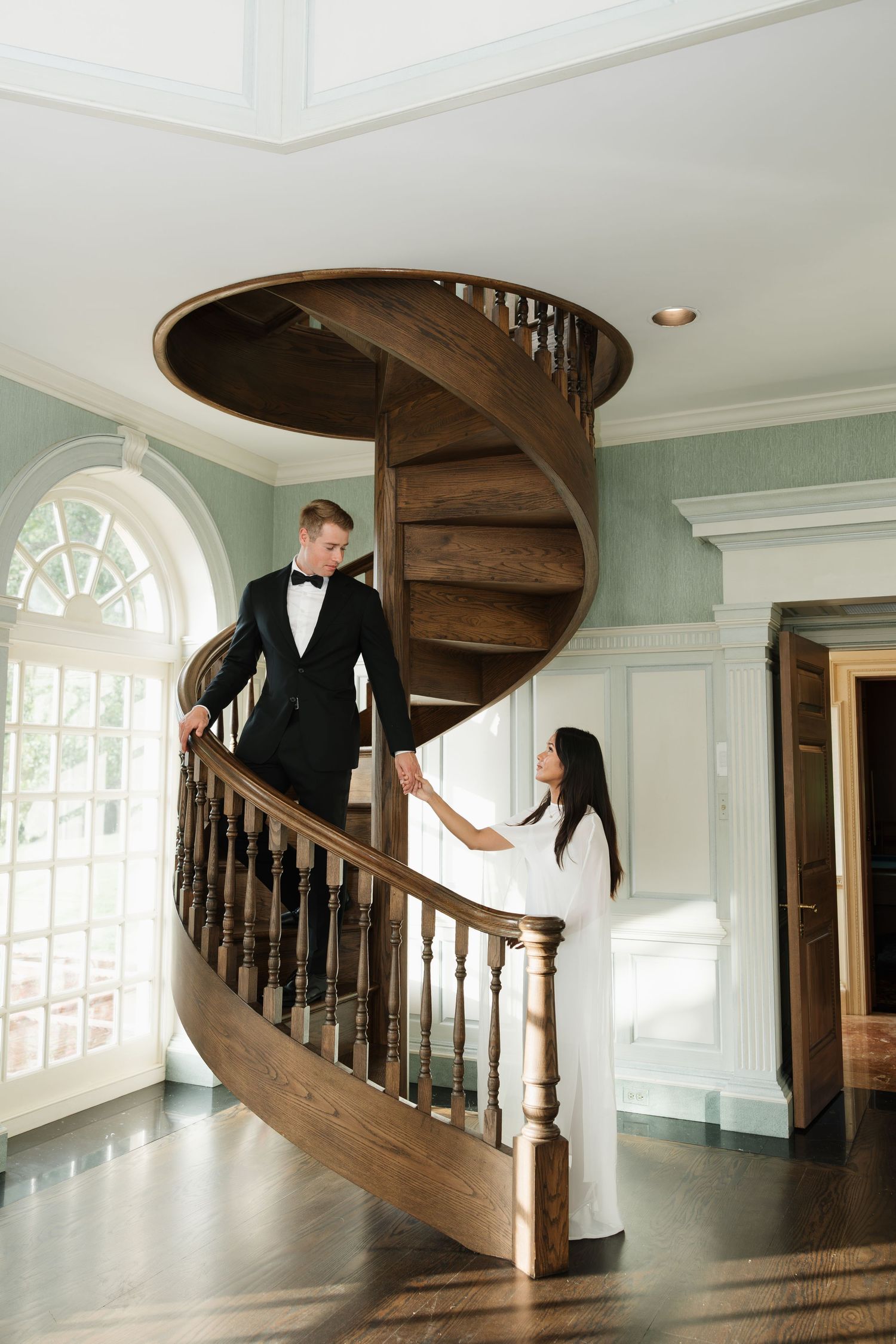 Couple in wedding attire holding hands on an elegant wooden spiral staircase in a classic manor home.