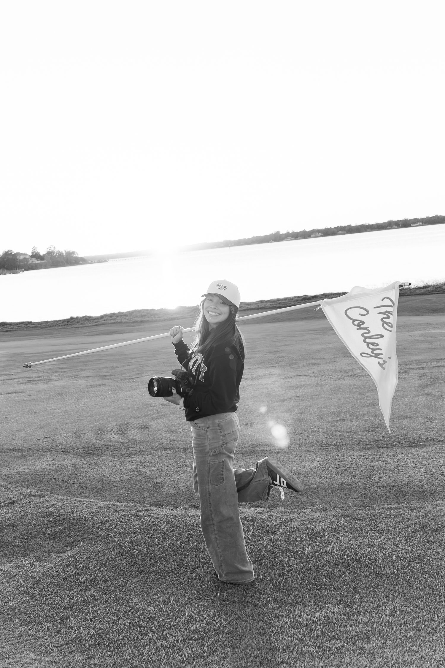 Black and white photo of a smiling woman holding a camera and a flag on a golf course near water.
