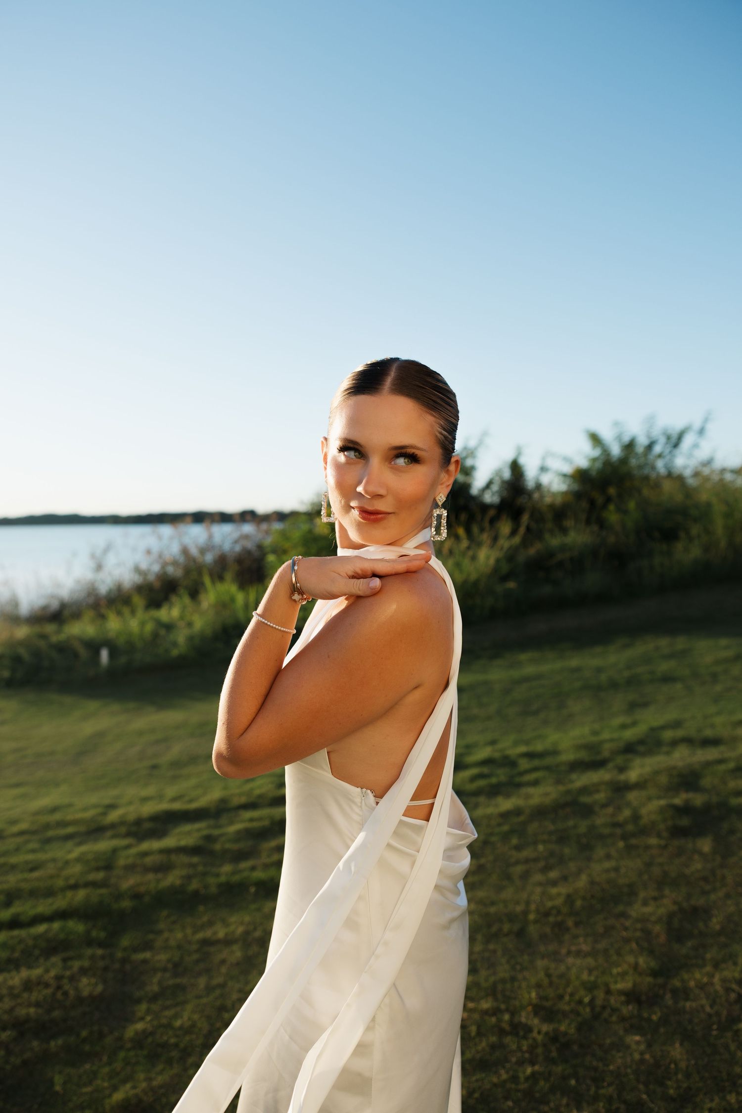 Elegant bride in white backless gown poses outdoors near waterfront at golden hour.