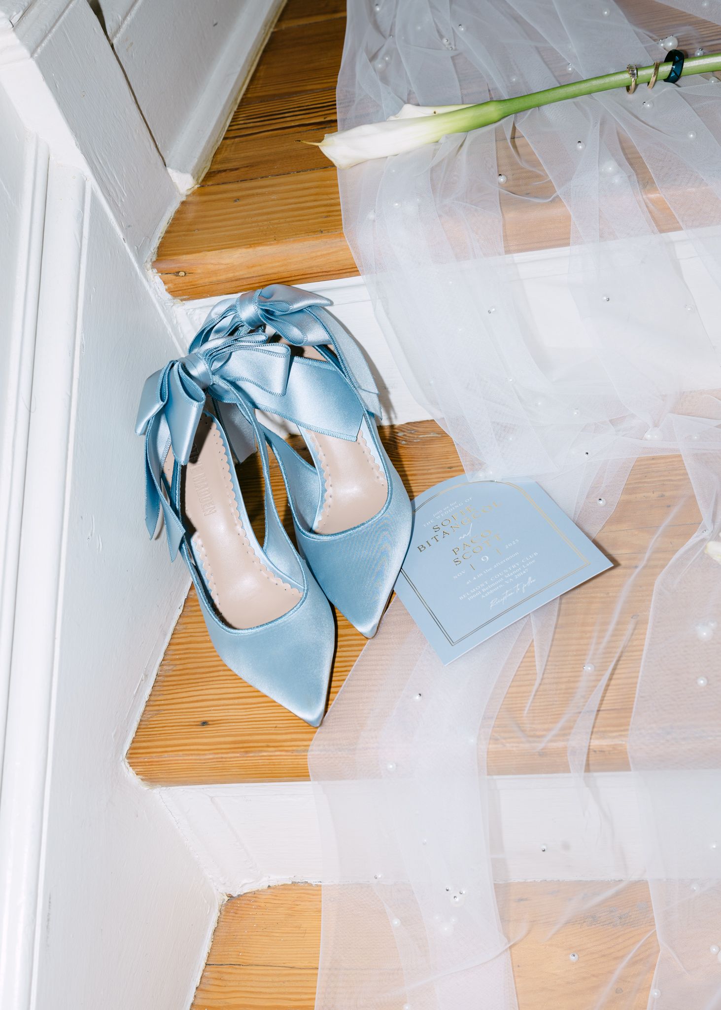 Light blue satin pointed-toe heels with bow ties beside wedding invitation and pearl-dotted veil on floor.