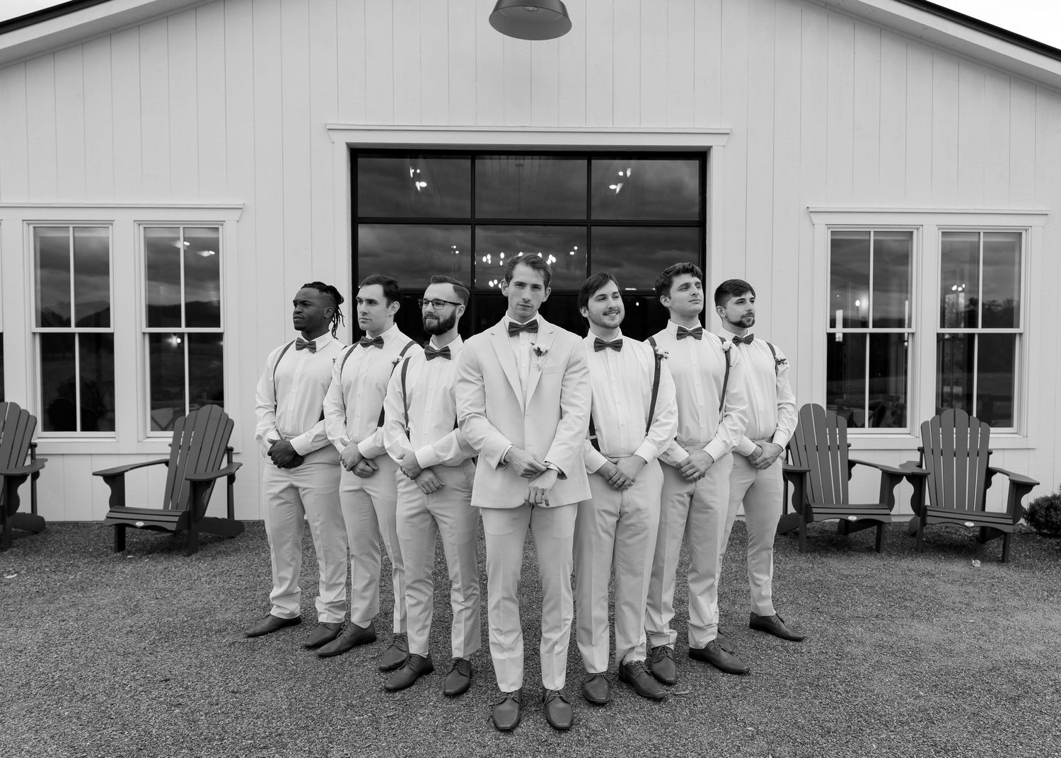 Groom and groomsmen in white suits and bow ties posing in front of a white barn.