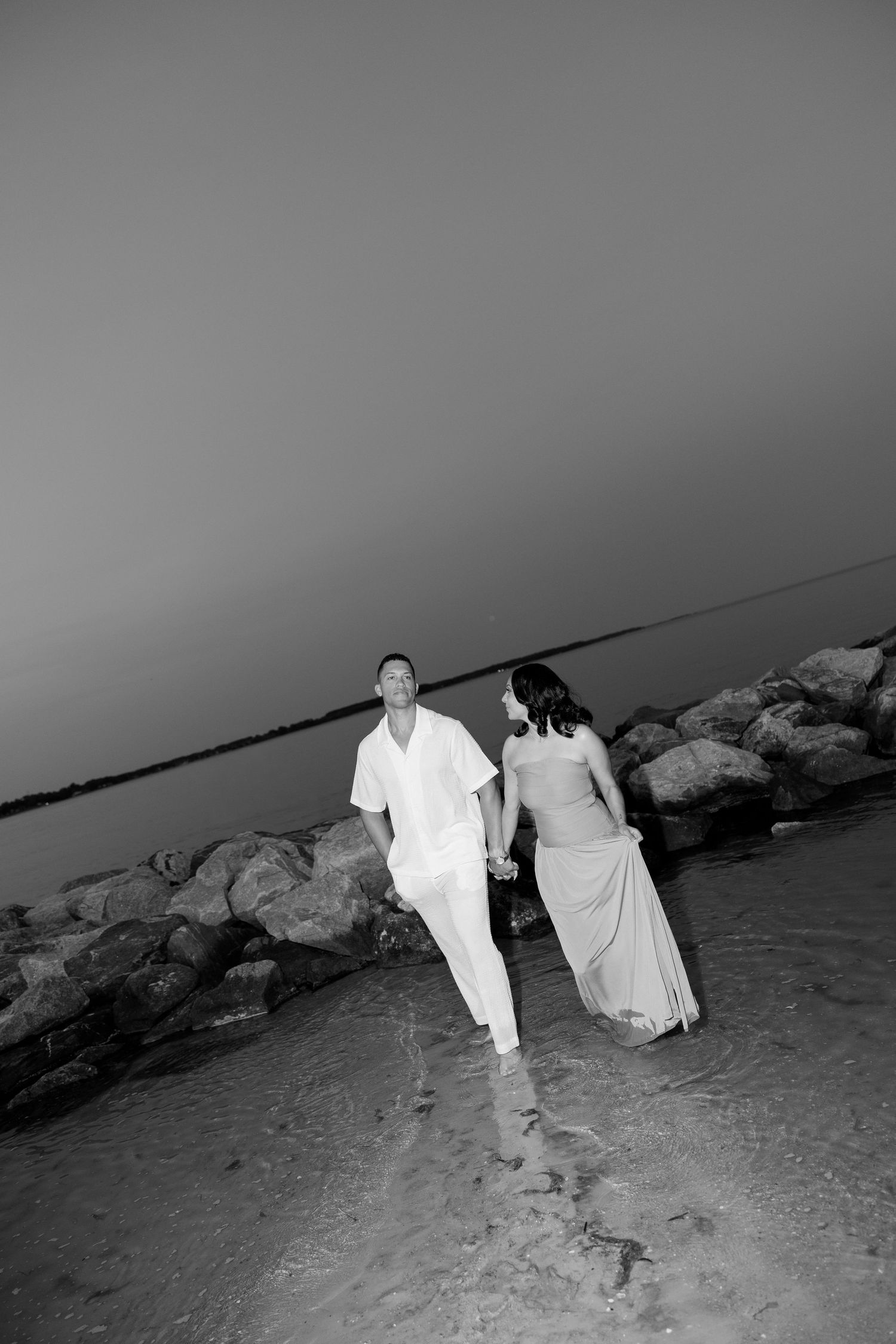 Black and white photo of a couple holding hands walking along rocky shoreline at sunset.