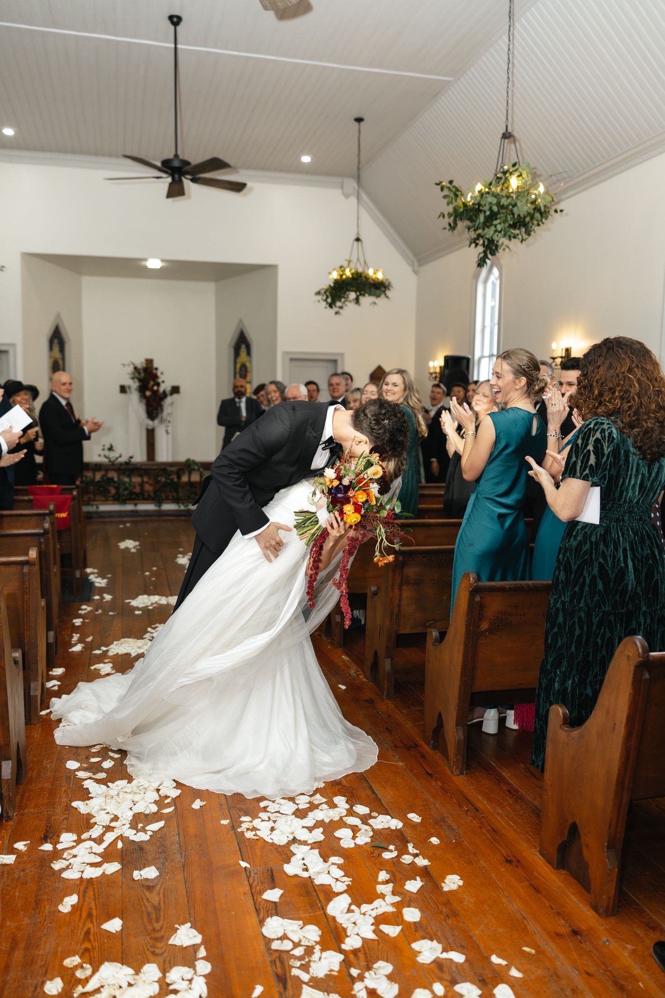 Newlyweds share a dip kiss while walking down the aisle as guests applaud in a charming white chapel.