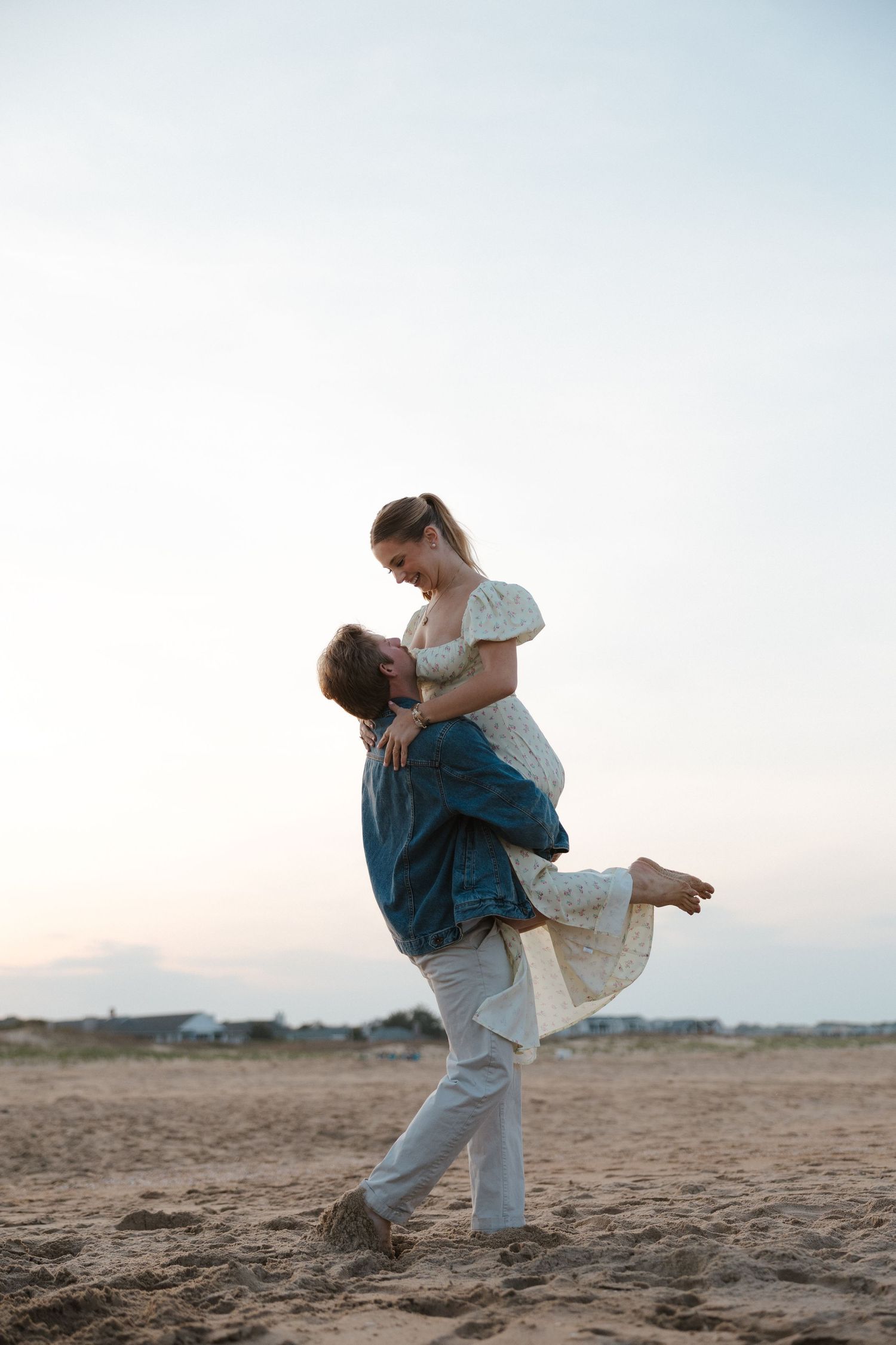 Man lifting woman in his arms on a sandy beach at dusk, both laughing joyfully.