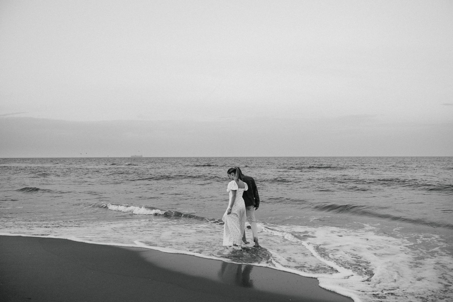 Couple shares romantic moment standing together at waters edge on beach in black and white.