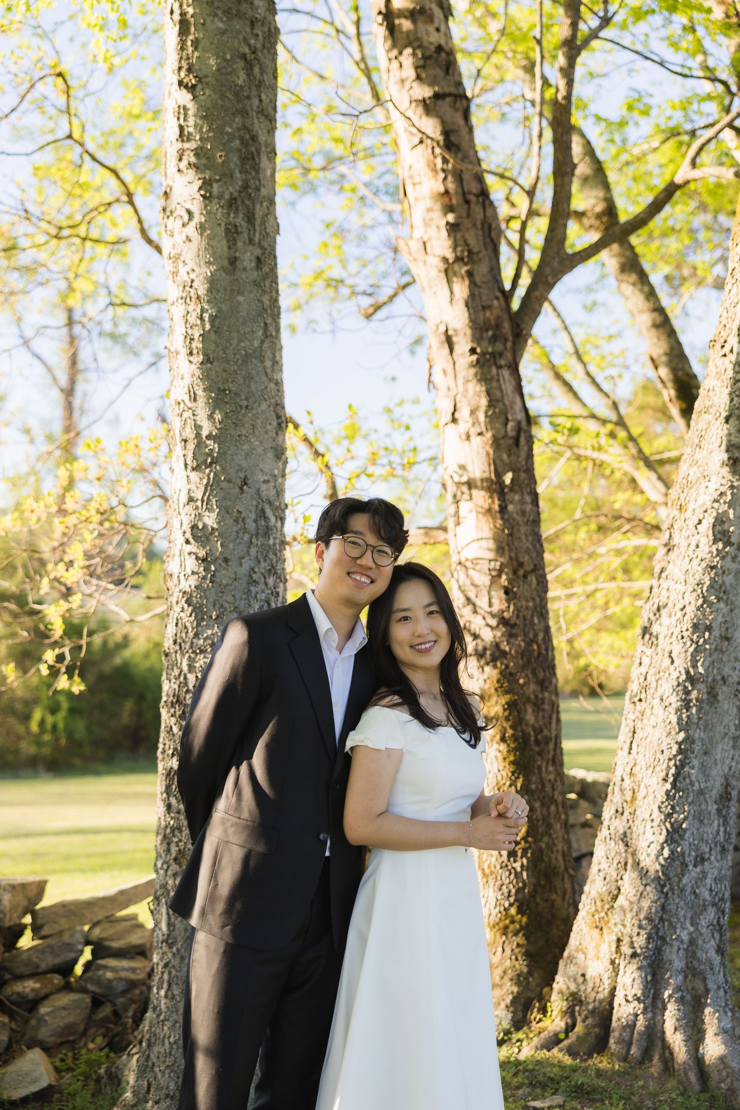 Smiling couple in wedding attire posing together between trees in a sunny outdoor park setting.
