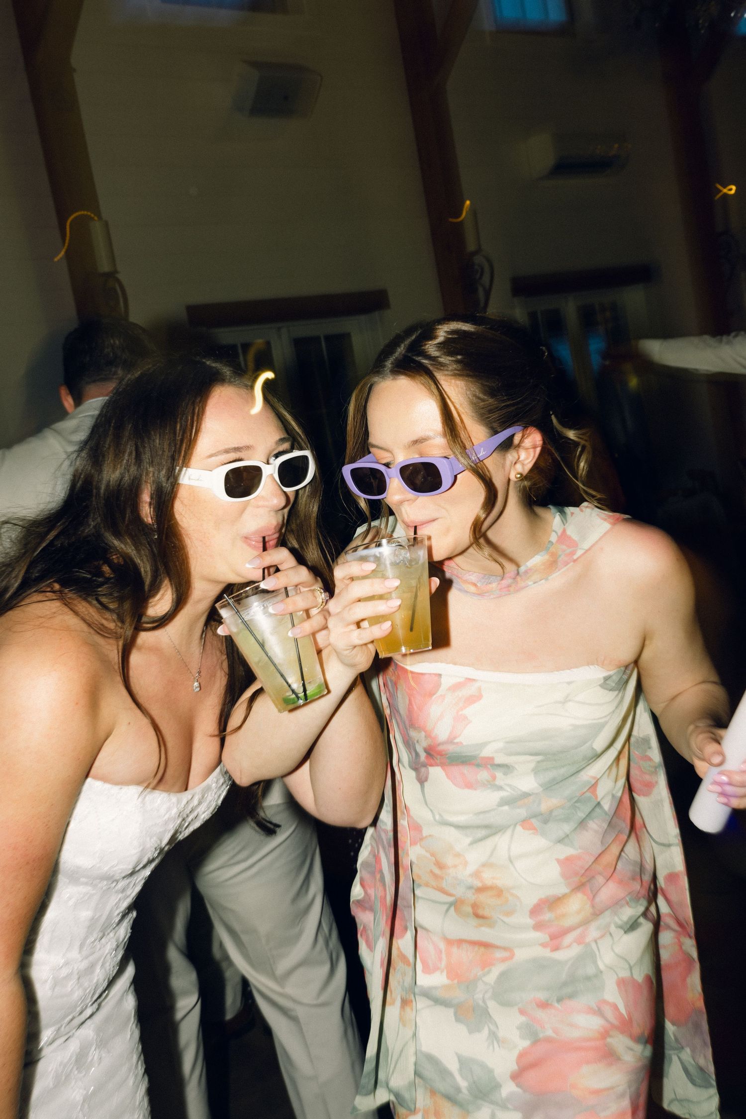 Two women in white dresses wearing sunglasses share drinks through straws at a wedding reception party.