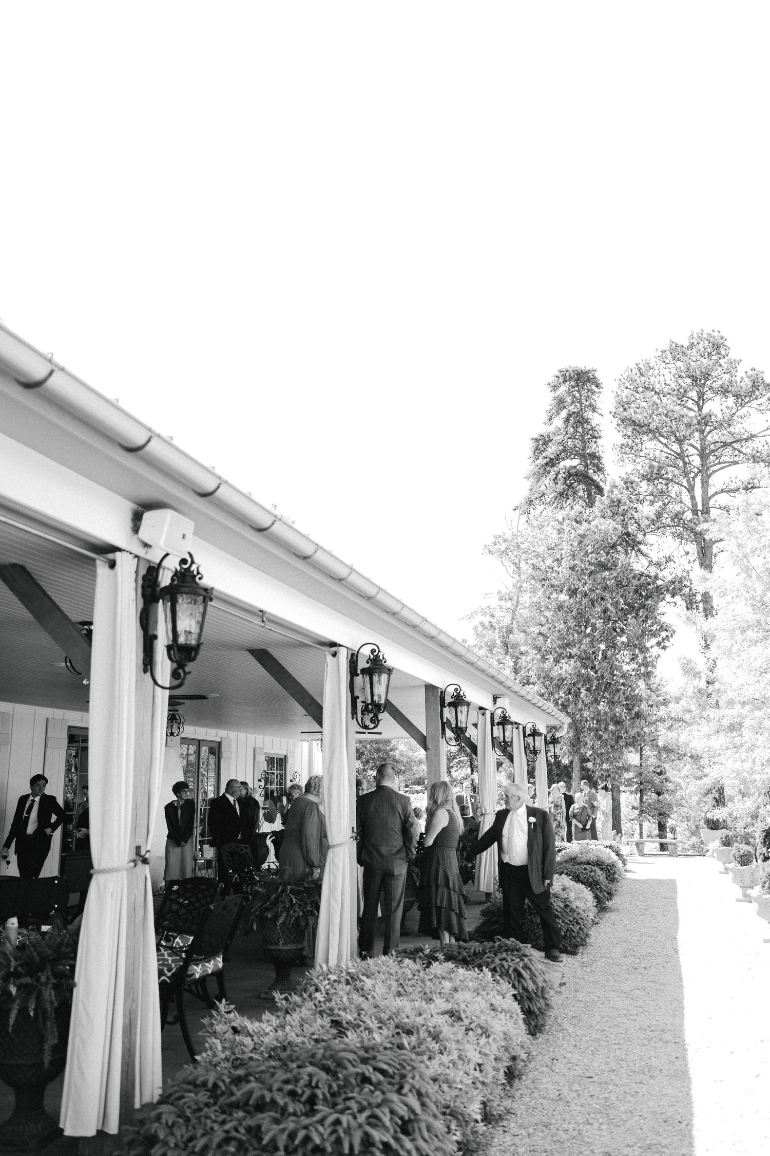 Black and white photo of elegantly dressed wedding guests mingling under a covered outdoor venue with lanterns.