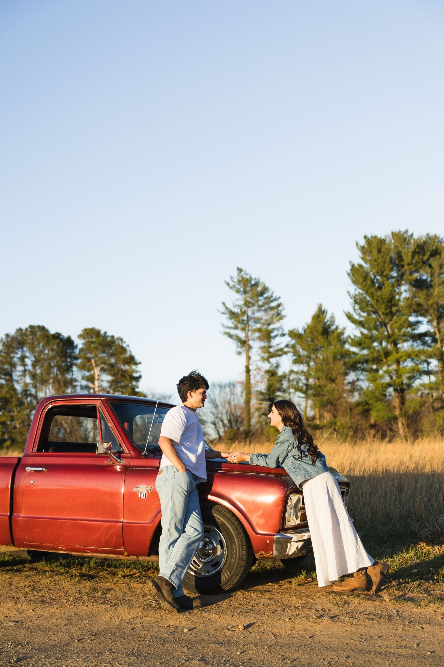 Young couple sharing a romantic moment leaning against a vintage red pickup truck in a rural field at golden hour.