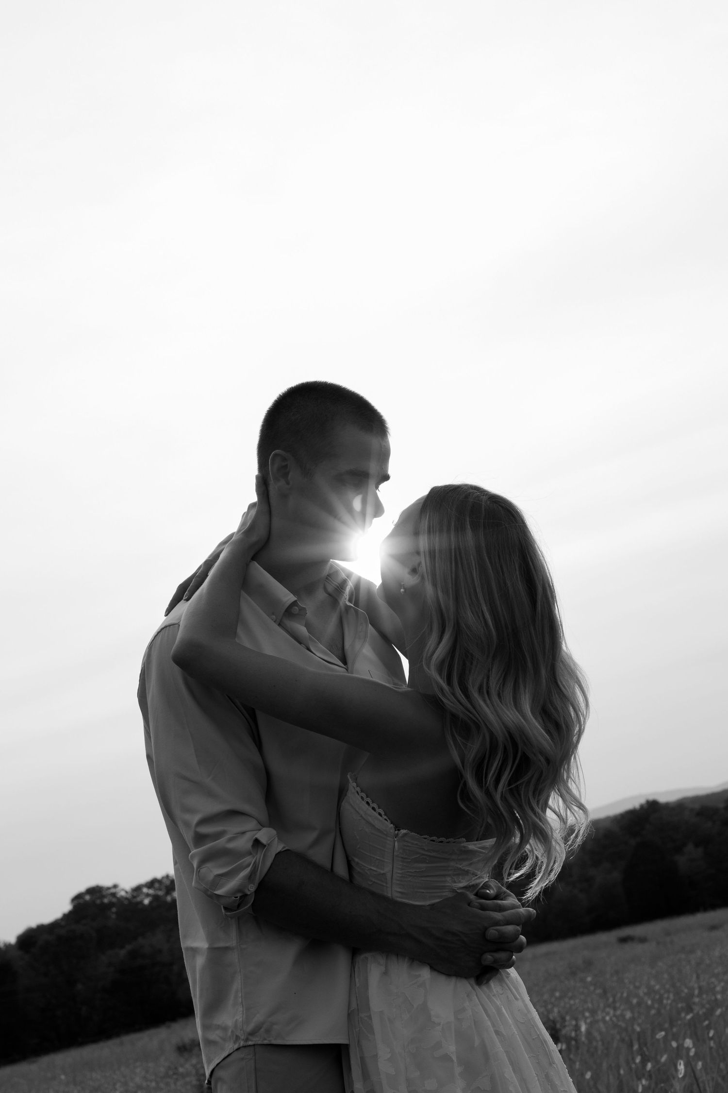 Black and white photo of a romantic couple embracing at sunset in a field, sun flare between them.