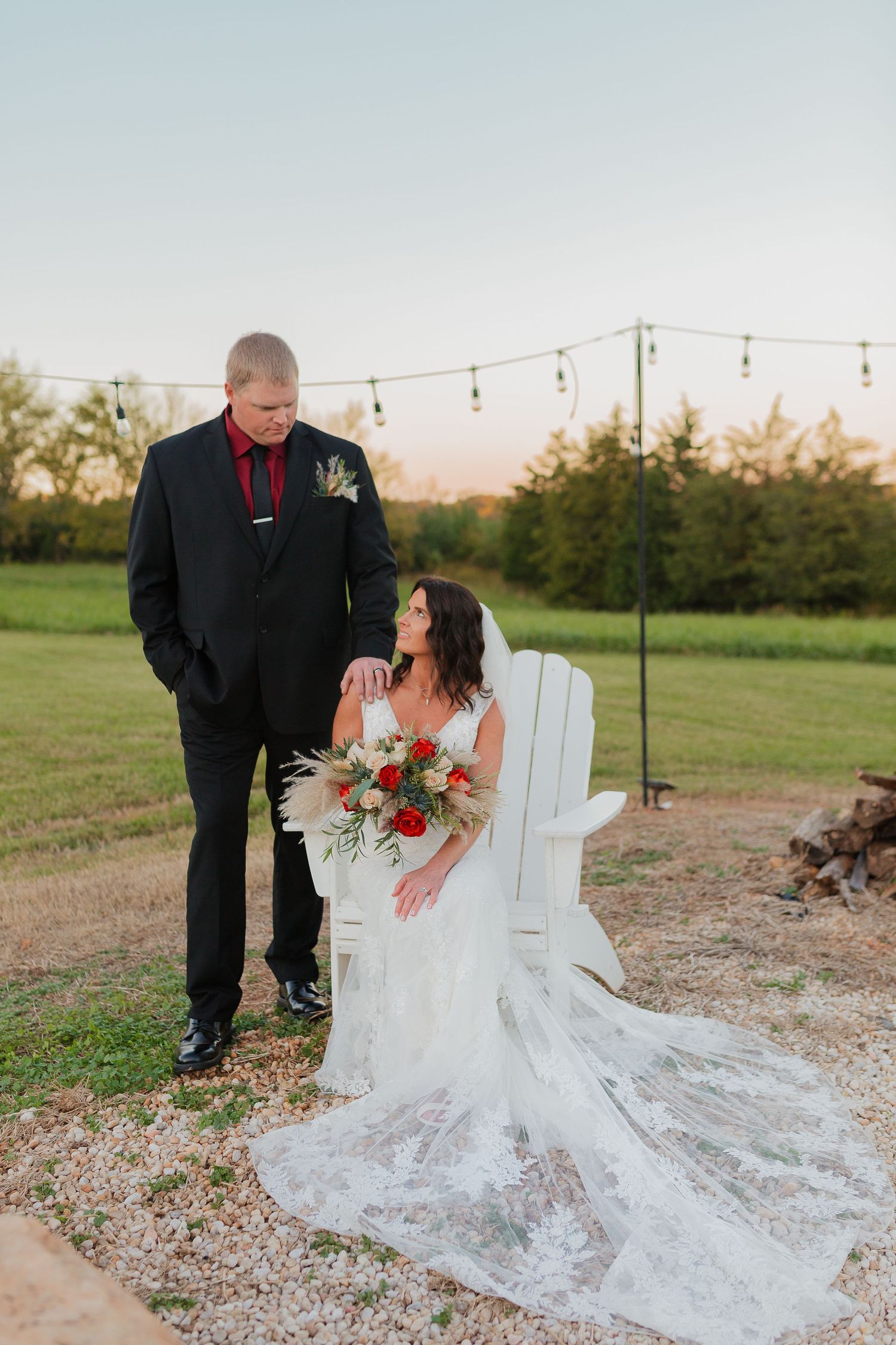 Bride holding red and white bouquet seated in white Adirondack chair with groom at golden hour outdoor wedding.