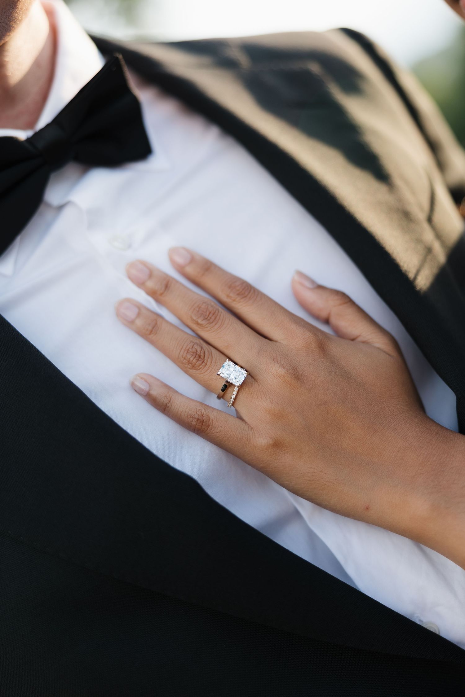 Close-up of a diamond engagement ring on a woman's hand resting on a man's tuxedo chest.