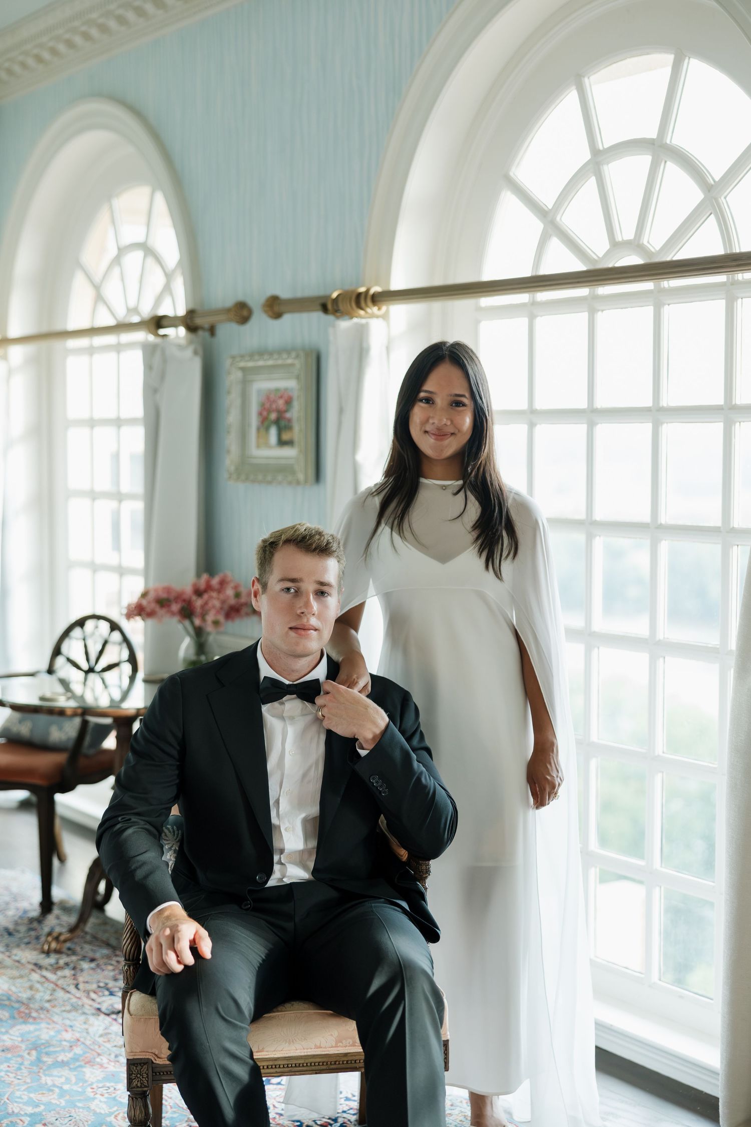 Elegant bride in white dress stands beside seated groom in black tuxedo in a bright, blue-walled room.