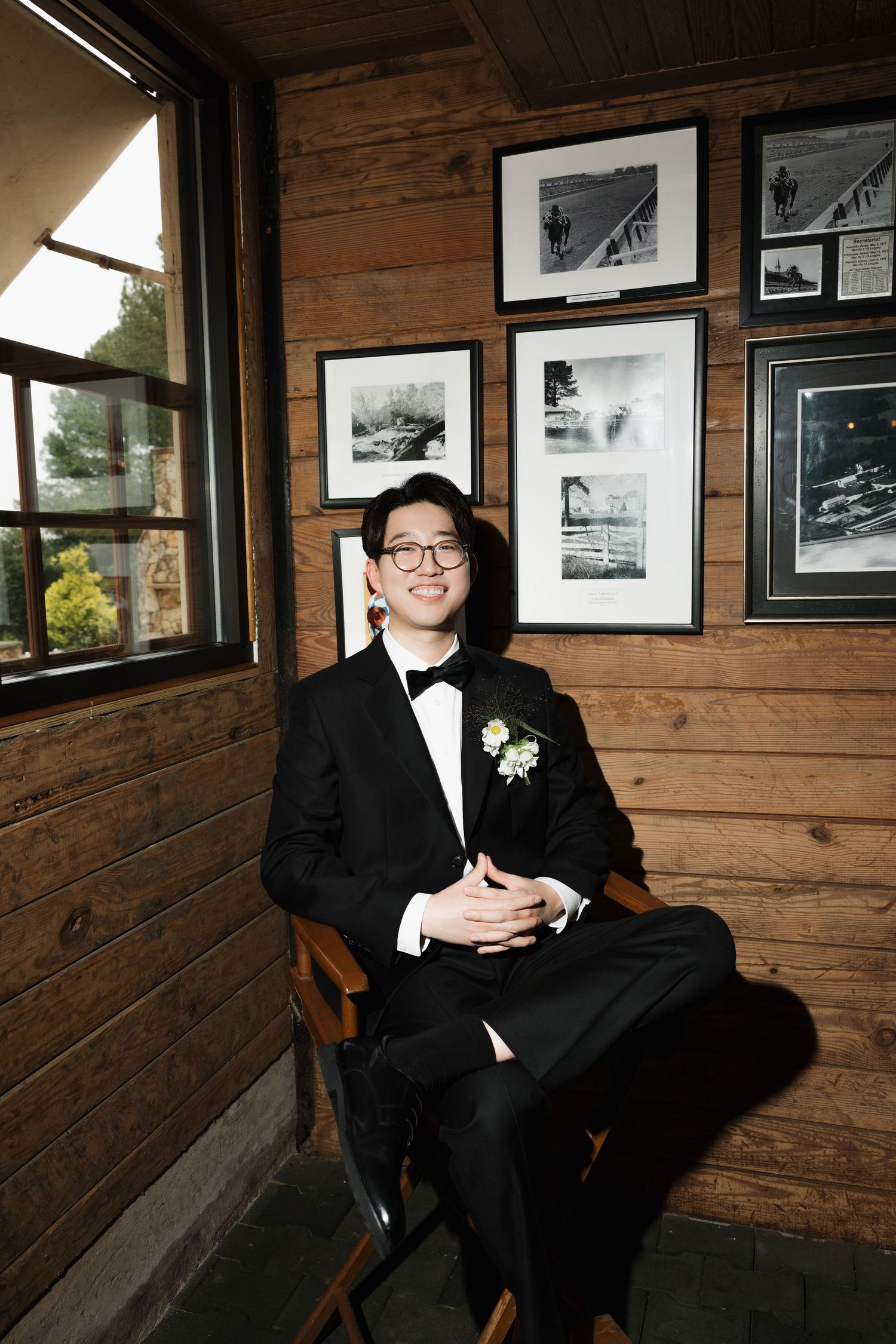 A young Asian man in a black tuxedo with boutonniere sits smiling in a wooden chair against a rustic wood-paneled wall.