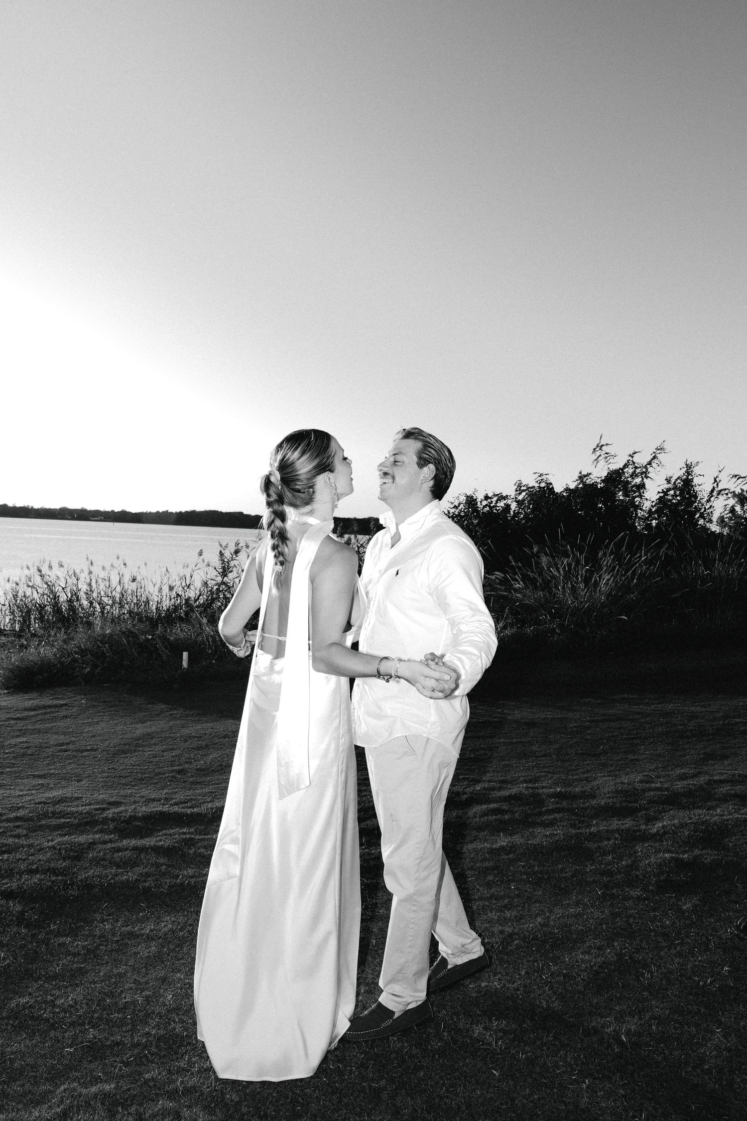 Black and white photo of a couple in white attire sharing a romantic moment by a waterfront at sunset.