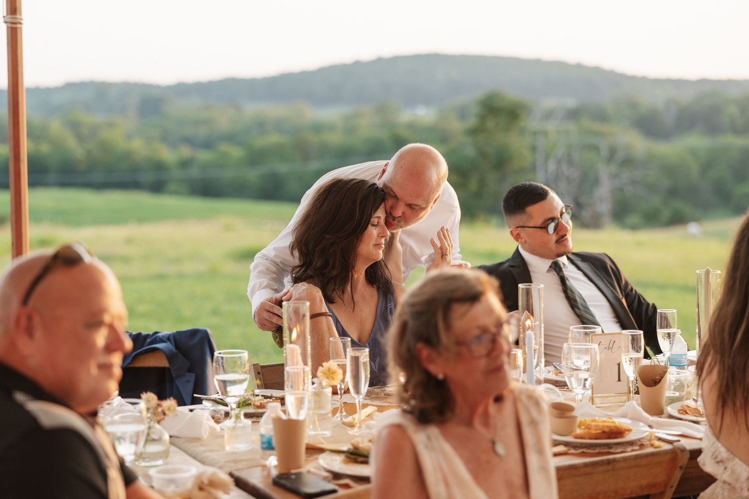 Guests socializing at an outdoor wedding reception dinner with scenic green hills in the background.