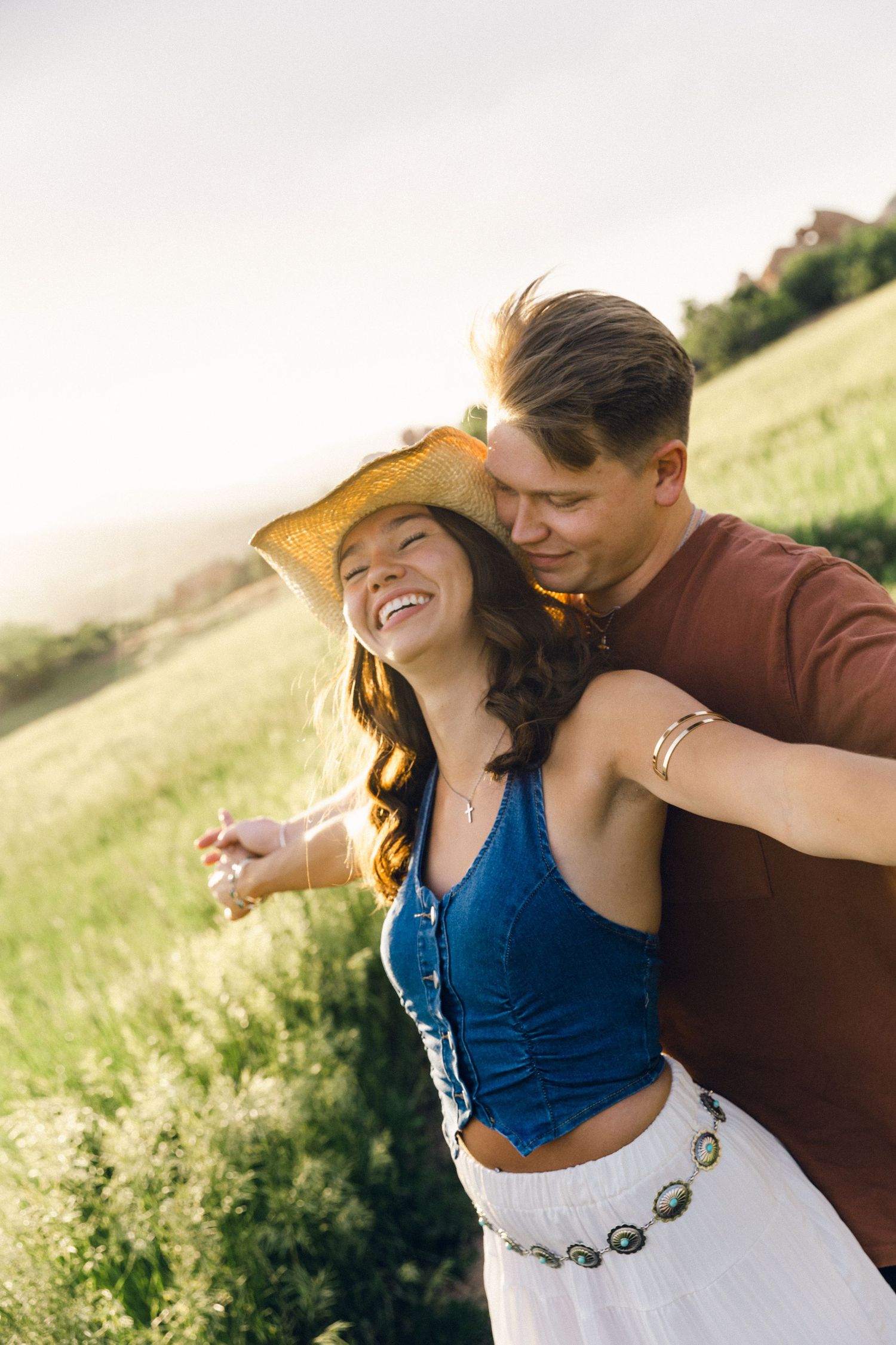 Couple laughing and embracing in a golden sunlit meadow, woman in cowboy hat and denim corset top.