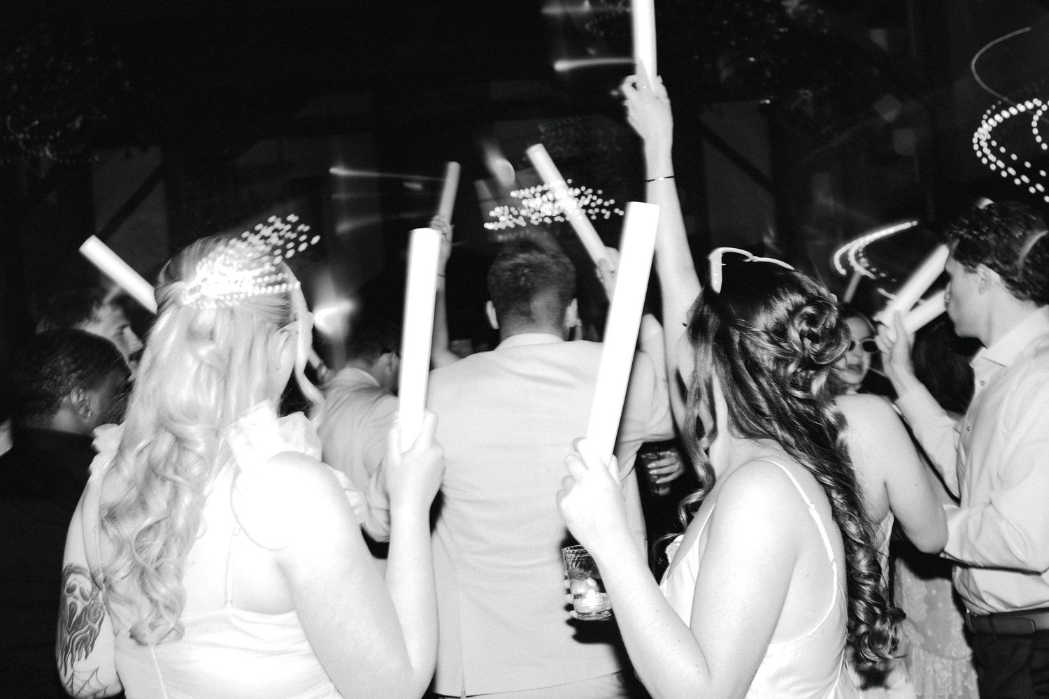 Black and white photo of wedding guests dancing with glow sticks at evening reception.