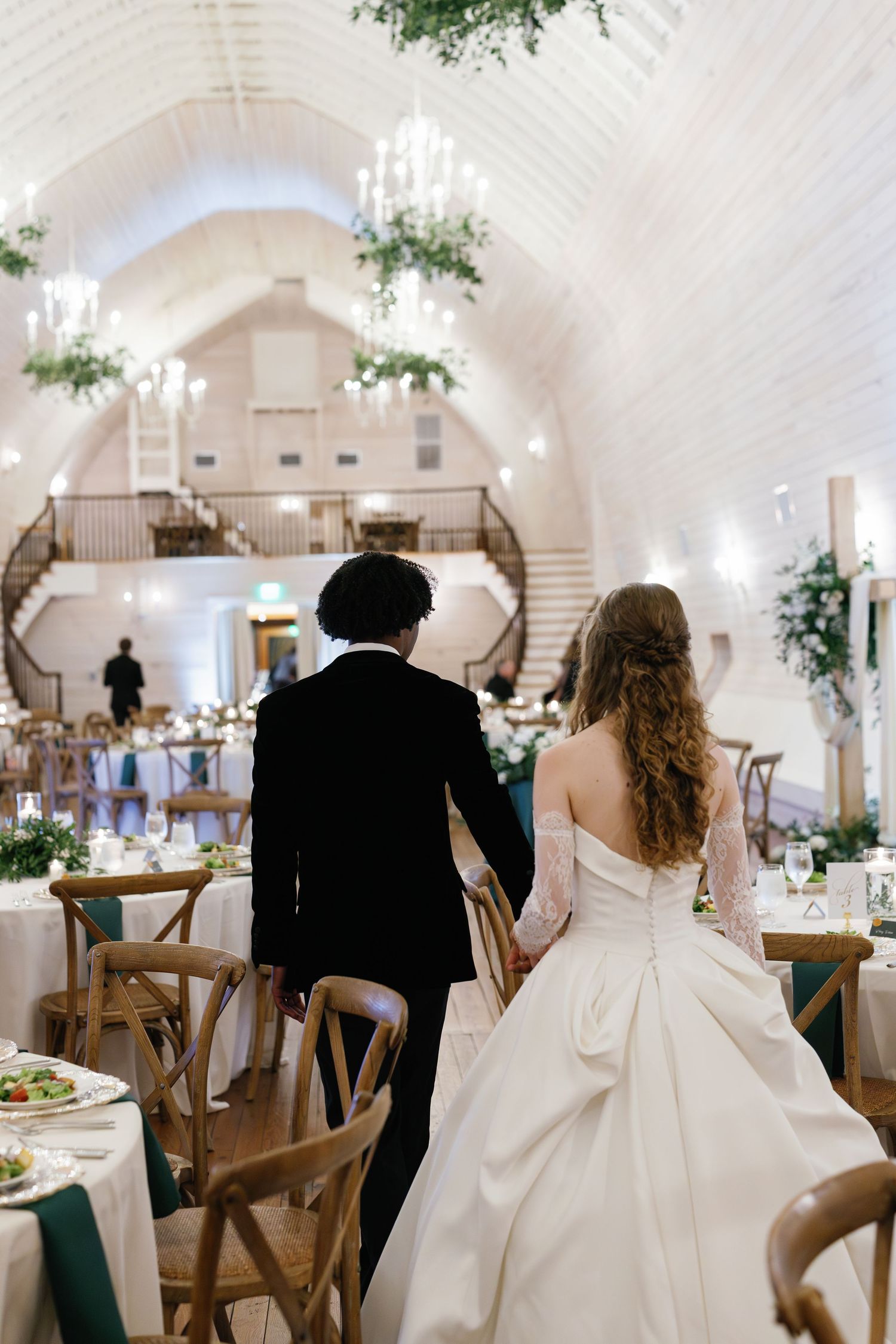 Bride and groom entering elegant white vaulted reception hall decorated with chandeliers and greenery.