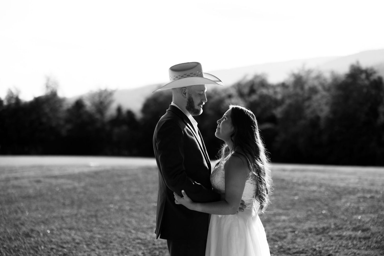 Black and white photo of a couple in wedding attire embracing in an open field with trees and mountains behind them.