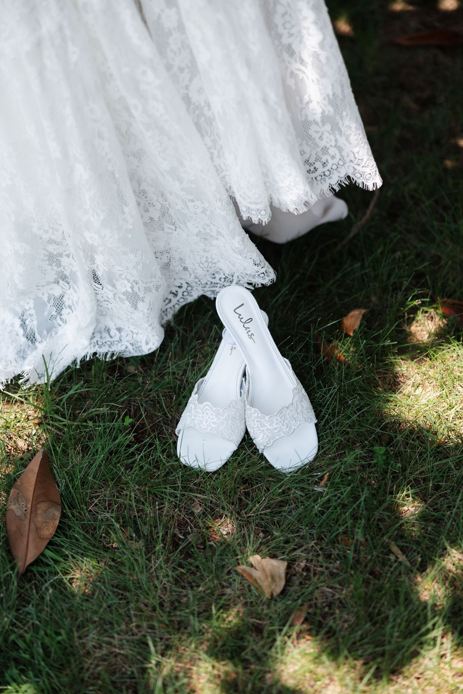 White lace wedding dress hem with beaded bridal heels on green grass.
