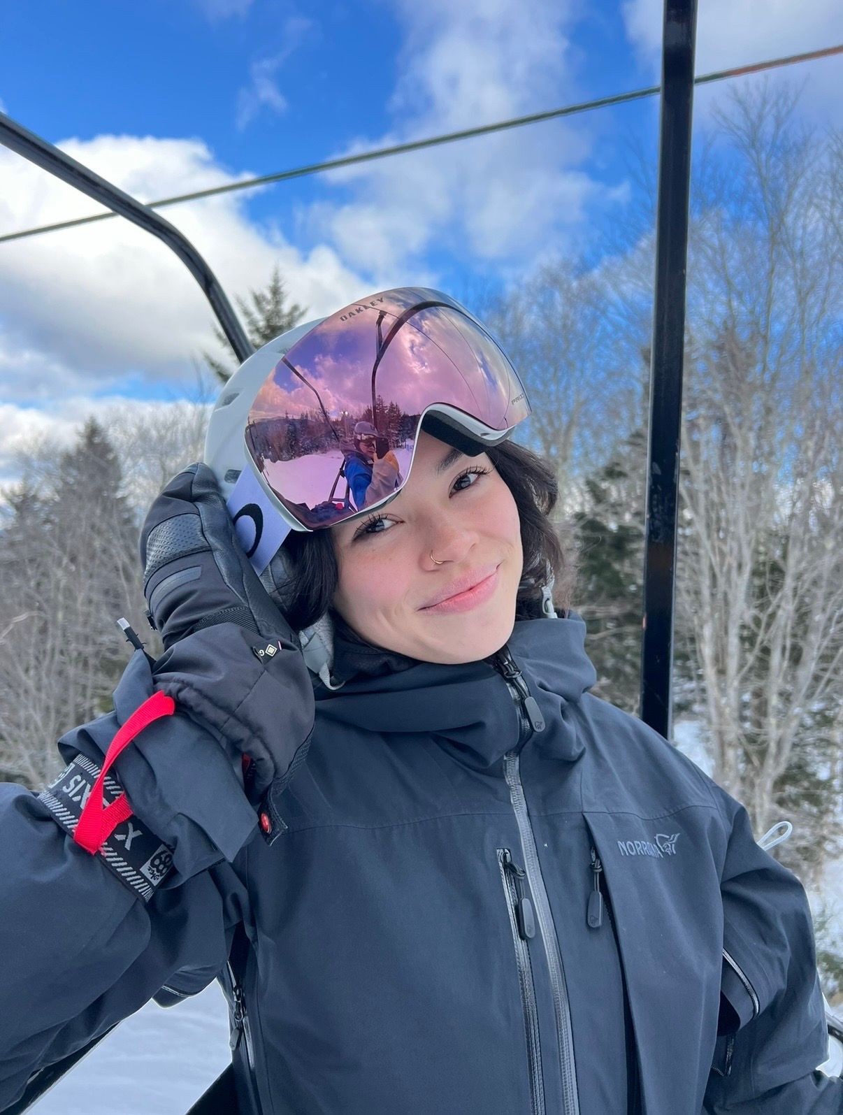 Young skier wearing pink goggles and black jacket on a ski lift on a sunny day.