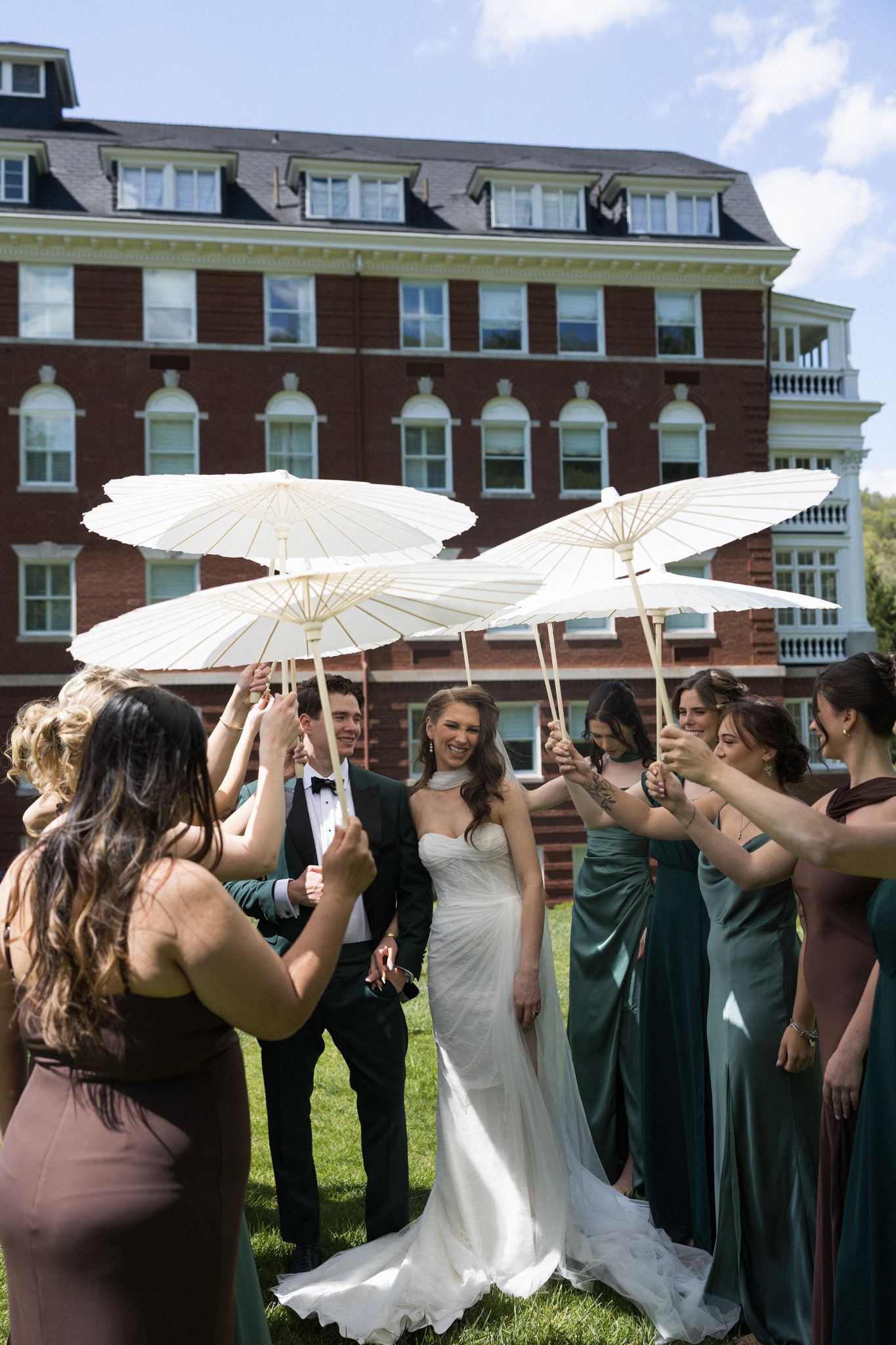 Wedding party holds white parasols over bride and groom outdoors near a red brick building on a sunny day.