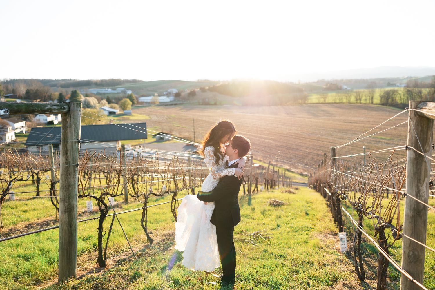 Bride and groom sharing a romantic embrace at golden hour in a scenic vineyard with rolling countryside views.