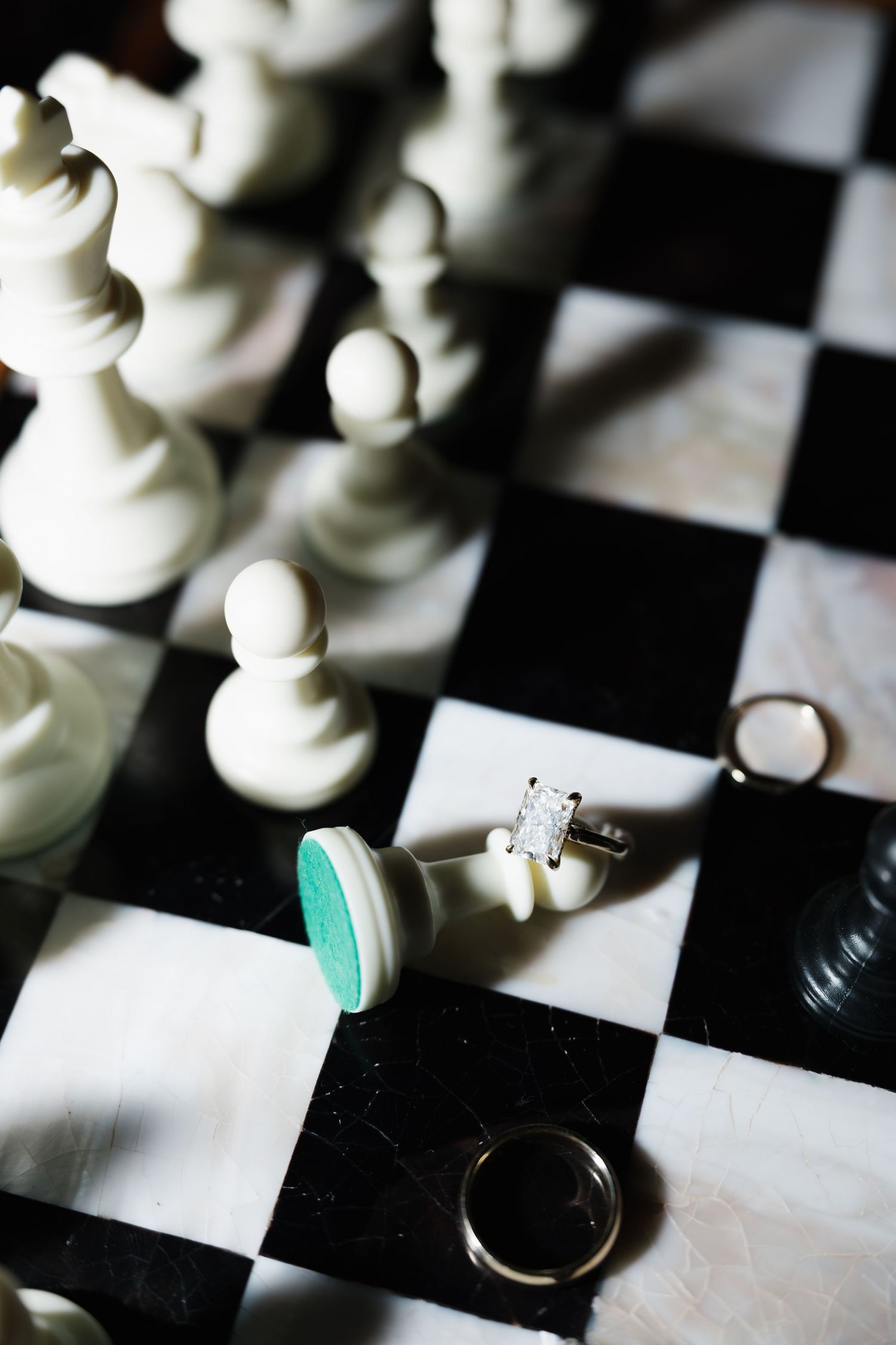 Diamond engagement ring and wedding bands displayed on a marble chess board surrounded by white chess pieces.