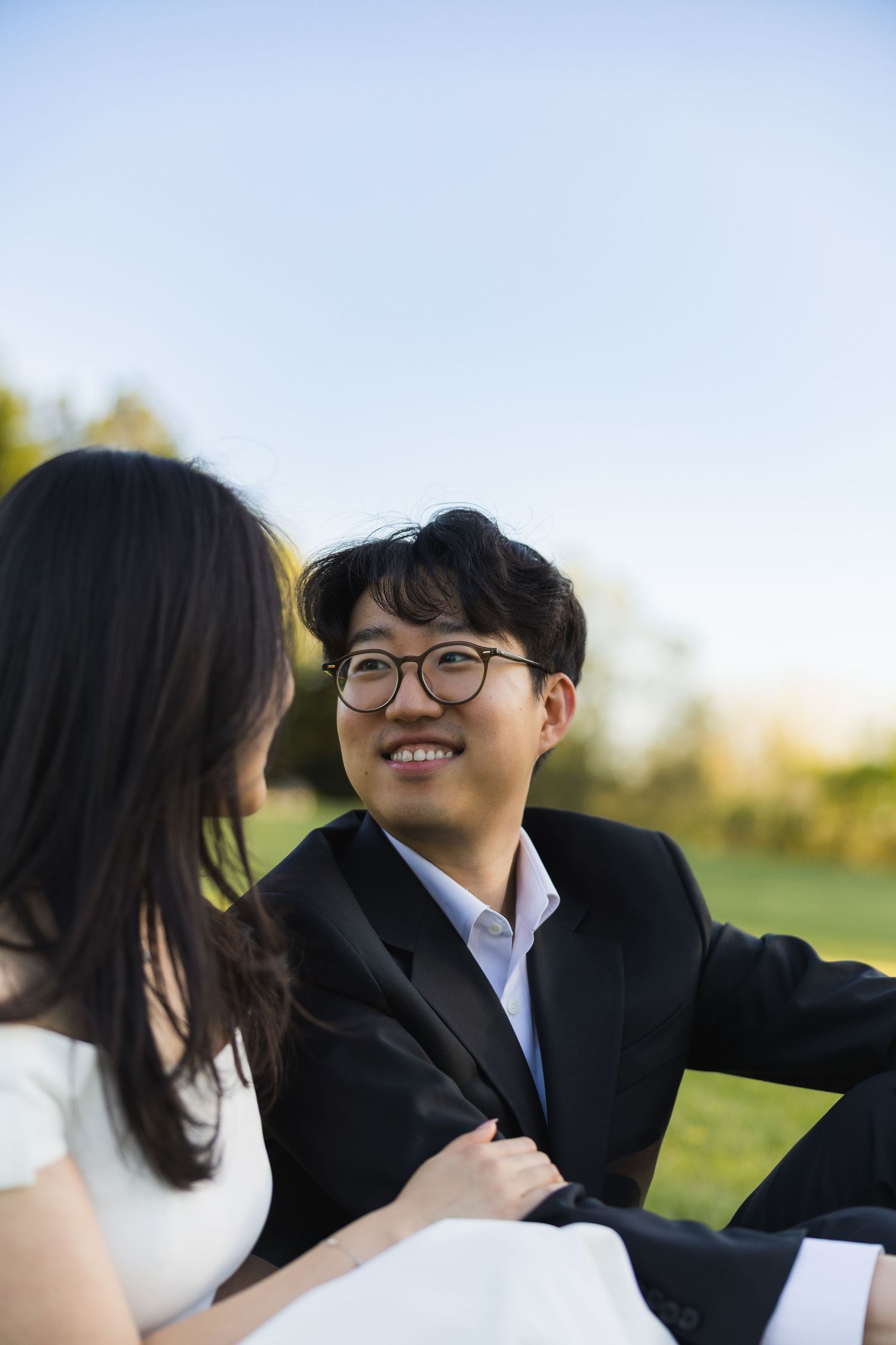Smiling groom in glasses and black suit looking at his bride while sitting on grass outdoors.